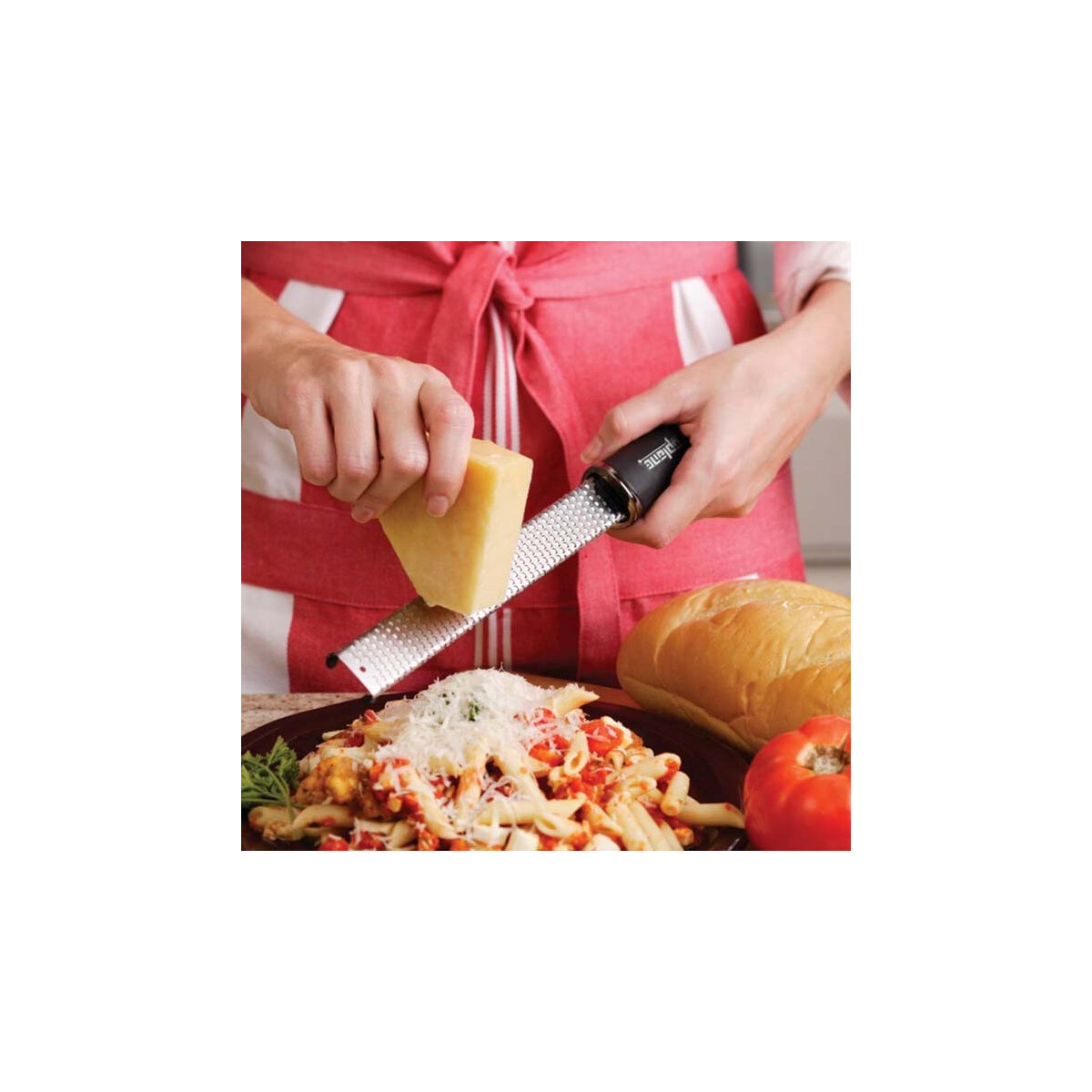 Eine Person reibt Käse über Pasta. Im Hintergrund ist eine rote Schürze. Brot und eine Tomate liegen daneben.