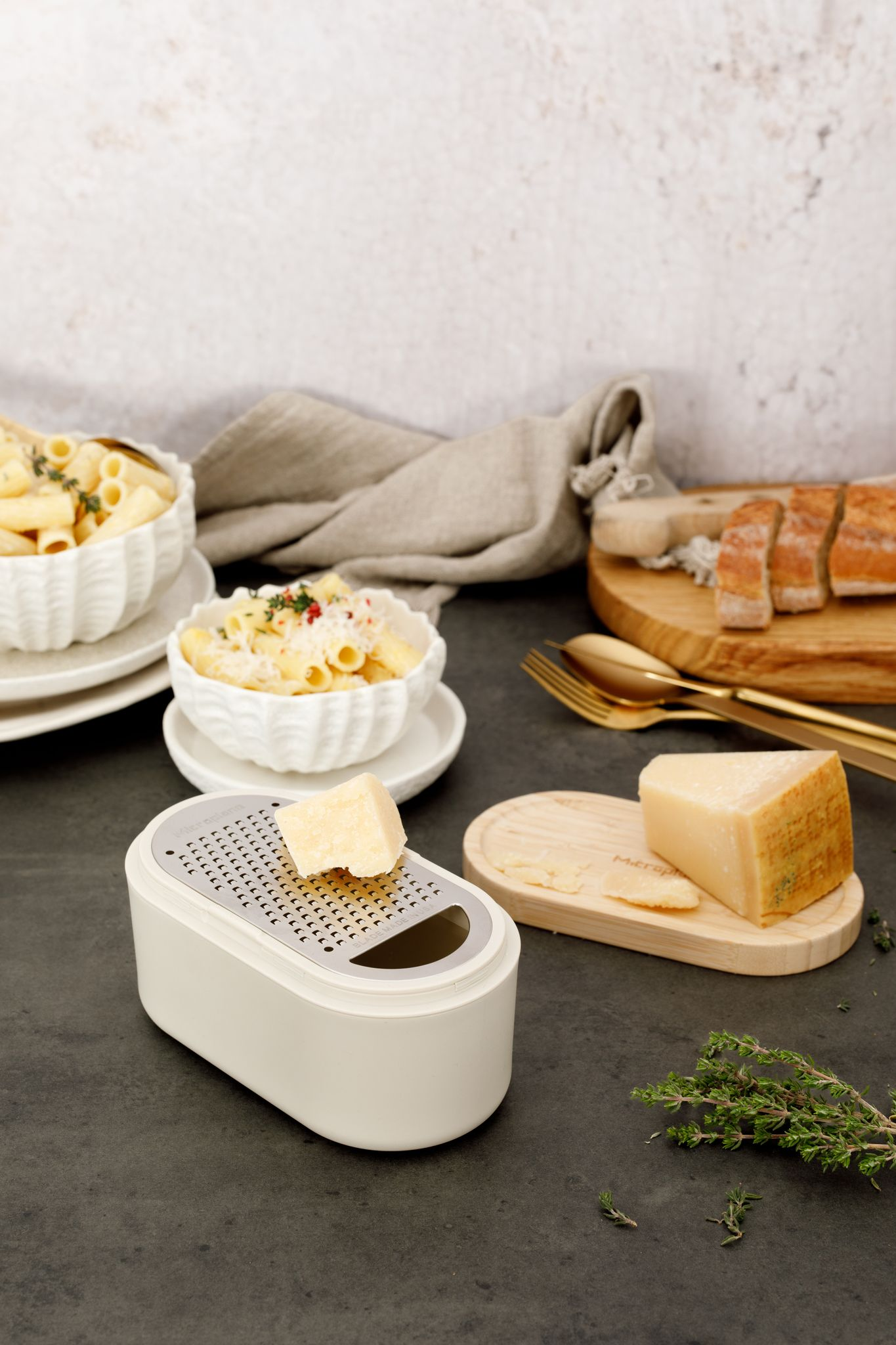 Pasta mit Käse in weißen Schalen, Käsehobel, Brot, Käsescheiben, goldenes Besteck, Kräuter auf dunkler Oberfläche.