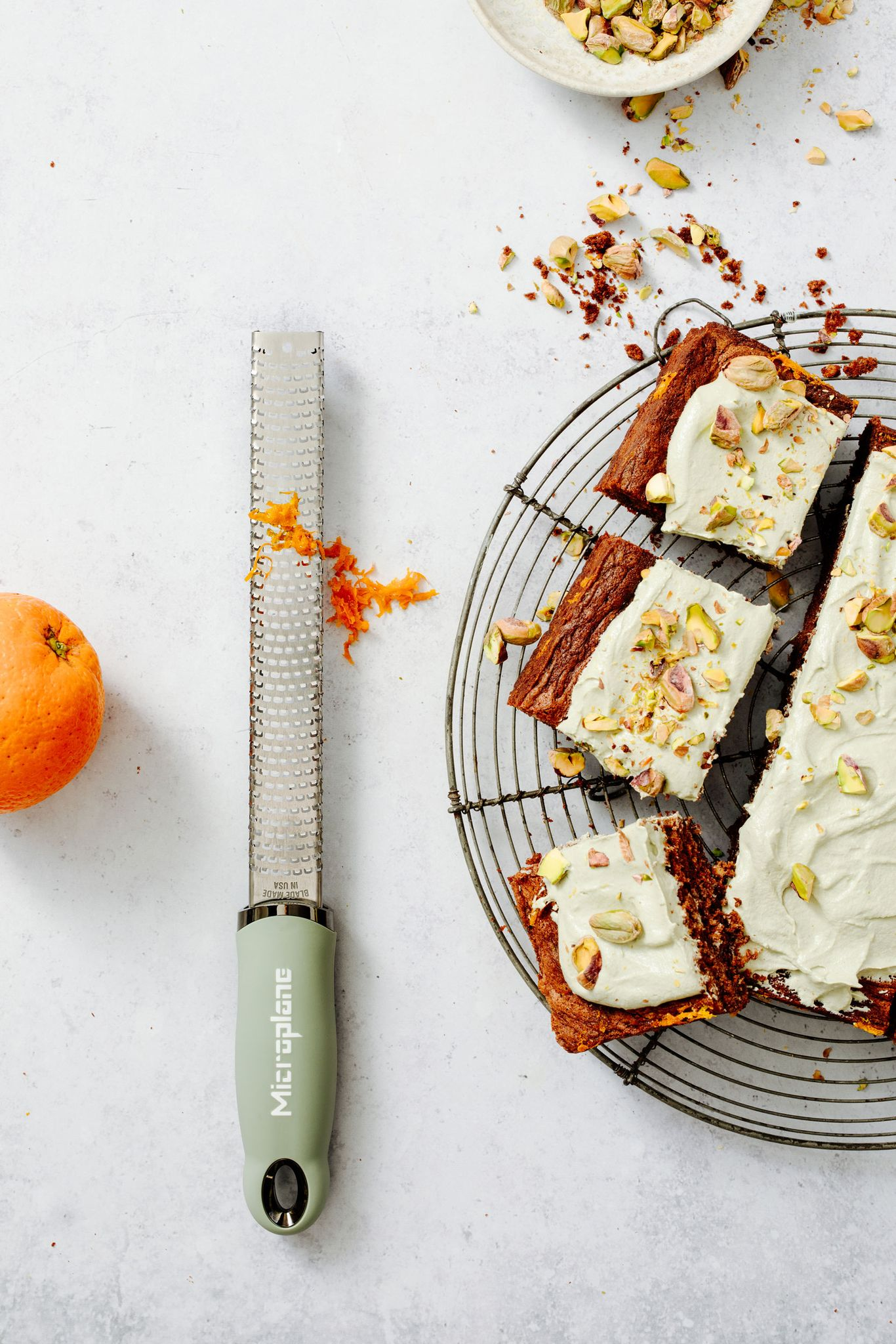 Draufsicht. Kuchenscheiben mit Zuckerguss und Pistazien auf einem Kühlgitter. Abrieb, Orange und Reibe liegen auf weißer Oberfläche.