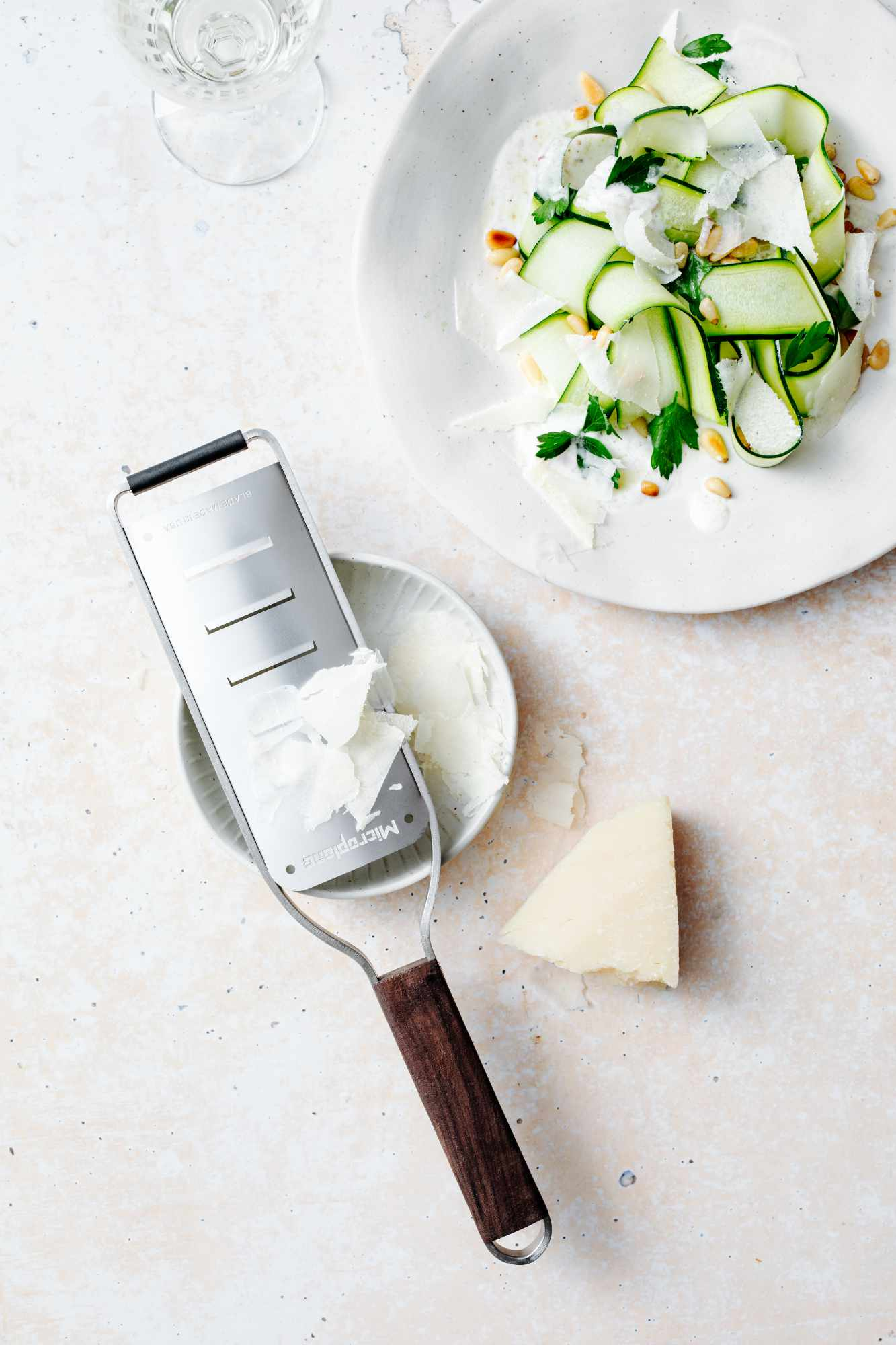Draufsicht auf Salat mit Zucchinibändern, Käse und Pinienkernen. Ein Reibeisen, Käse und ein Glas sind ebenfalls vorhanden.