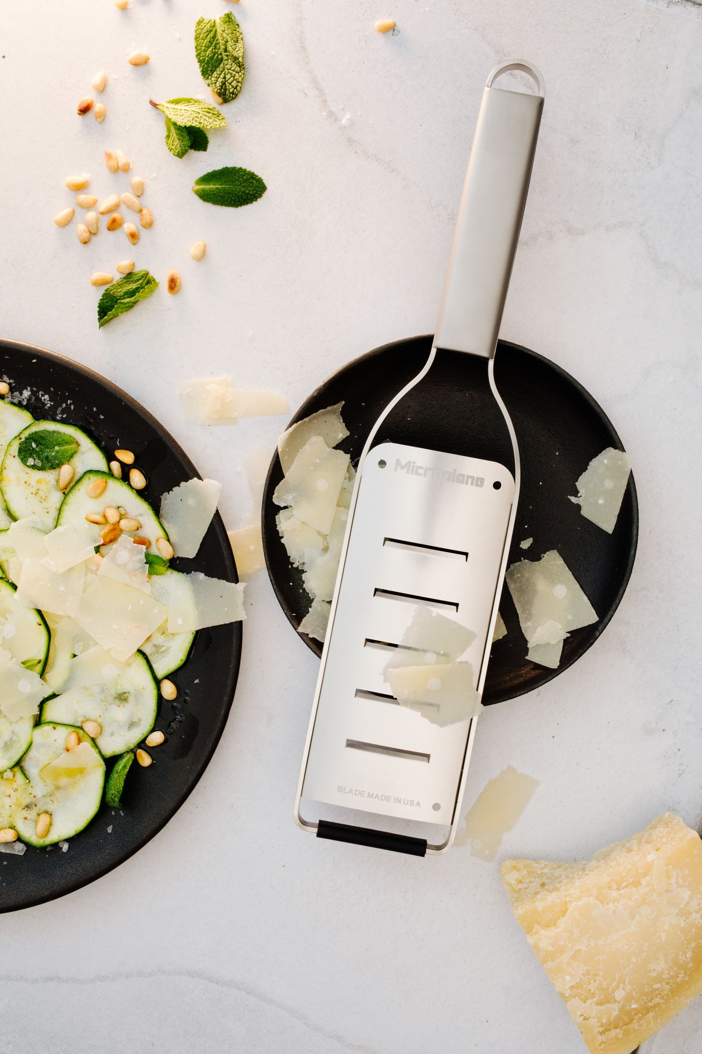 Draufsicht auf eine Reibe, Käse und geschnittene Zucchini auf schwarzen Tellern. Pinienkerne und Minzblätter sind verstreut.