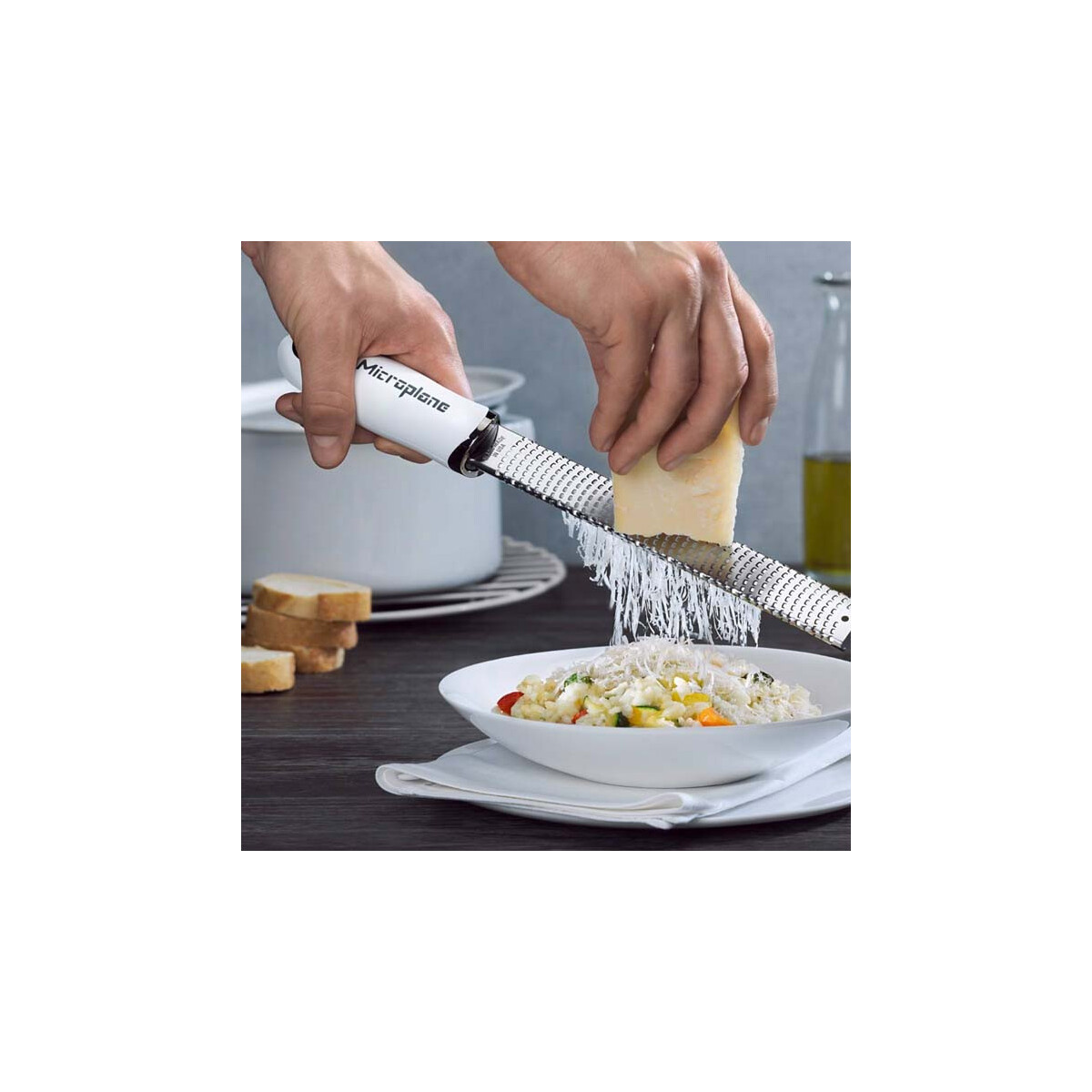 Hände reiben Käse über eine Schüssel Risotto. Ein weißer Reibeisen mit dem Wort 'Microplane' wird verwendet. Zutaten sind Gemüse und Käse.