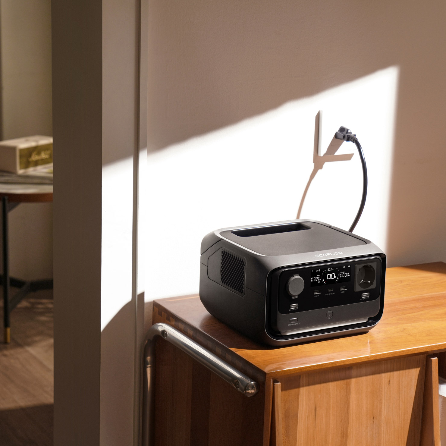 Czarno-szara stacja zasilania EcoFlow na drewnianym stole. Czarny kabel jest podłączony do gniazdka ściennego. Tło to biała ściana.