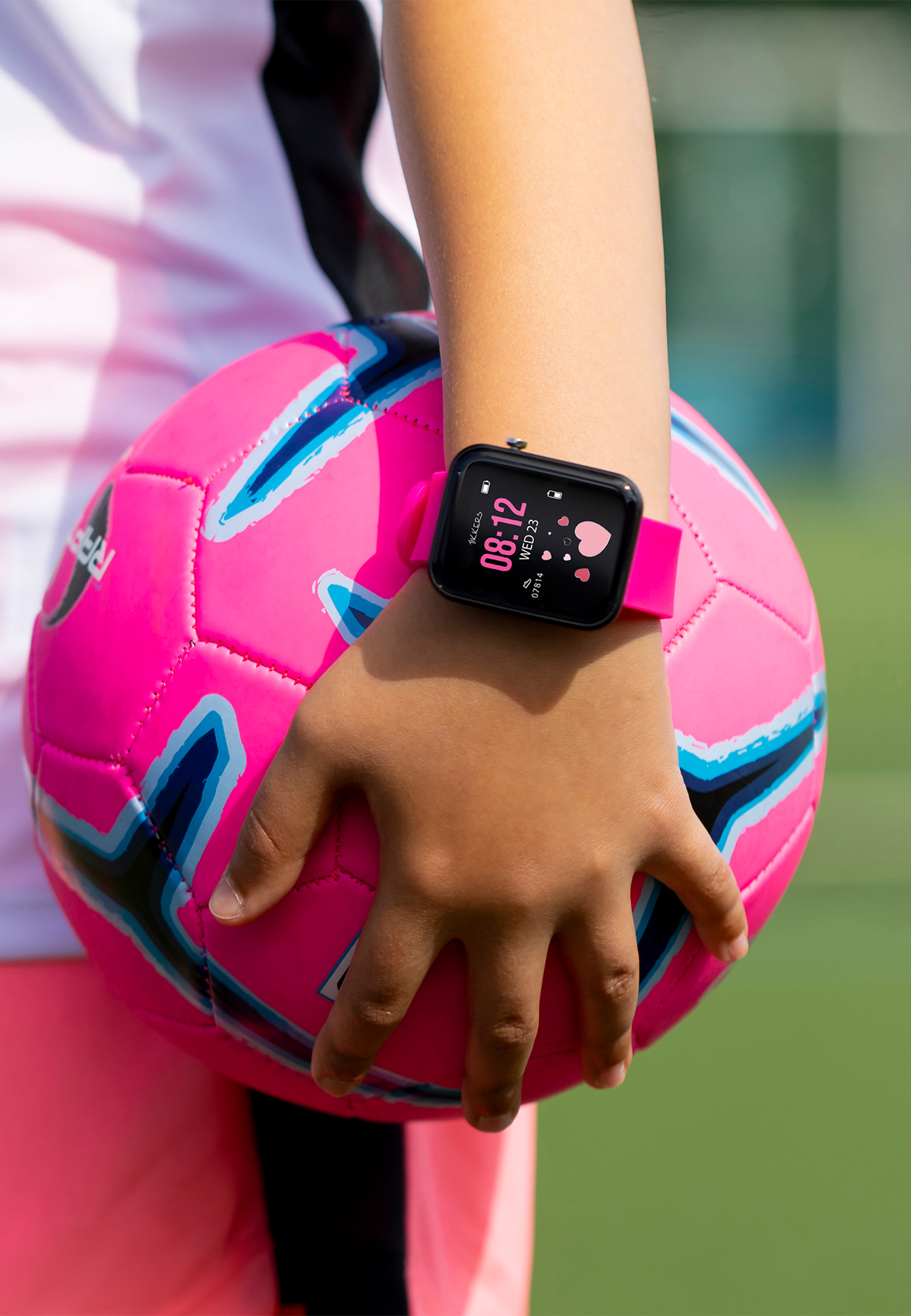 Eine Person hält einen pinkfarbenen Fußball. Eine Uhr am Handgelenk zeigt die Zeit und ein Herzsymbol. Grüner Hintergrund.