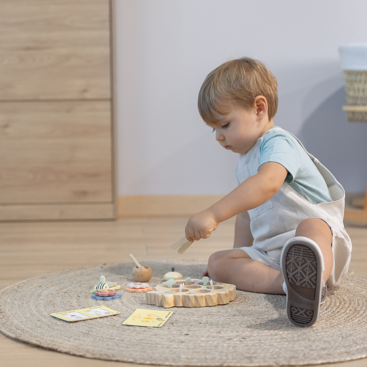 Un niño pequeño juega con un juguete de madera sobre una alfombra. Hay cartas y otras piezas de madera cerca. Viste ropa clara y está concentrado.