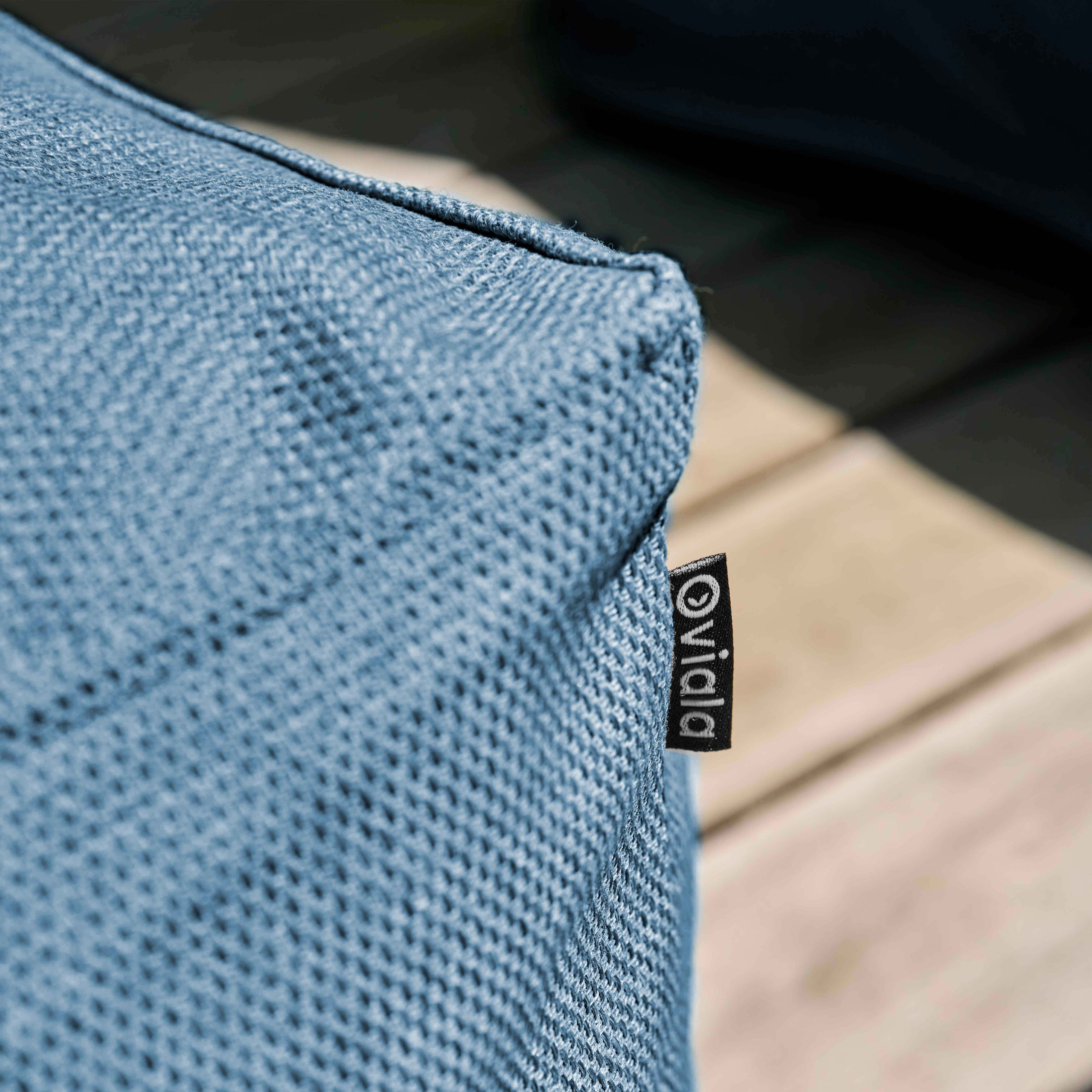 Nahaufnahme eines blauen Kissens. Ein schwarzes Etikett mit weißem Text 'Vivida' ist angebracht. Hintergrund zeigt eine Terrasse.