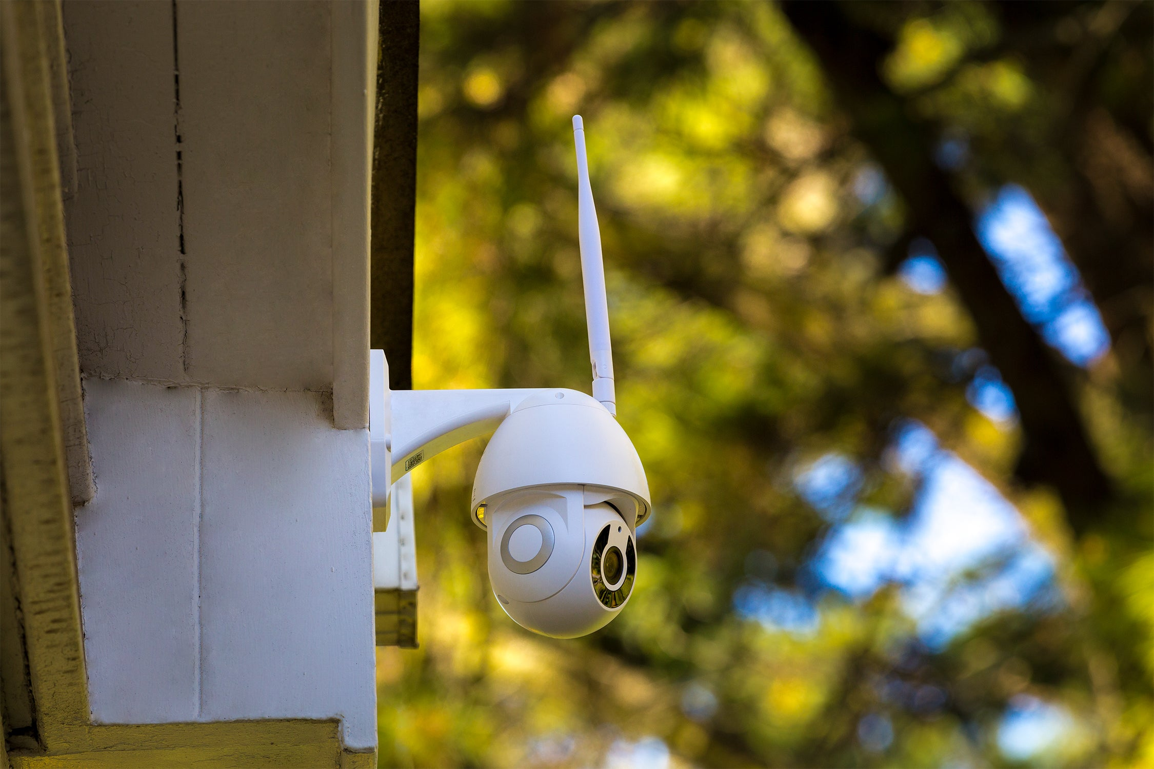 Cámara de seguridad blanca montada en una esquina de un edificio, con antena blanca. Follaje verde y amarillo en el fondo.