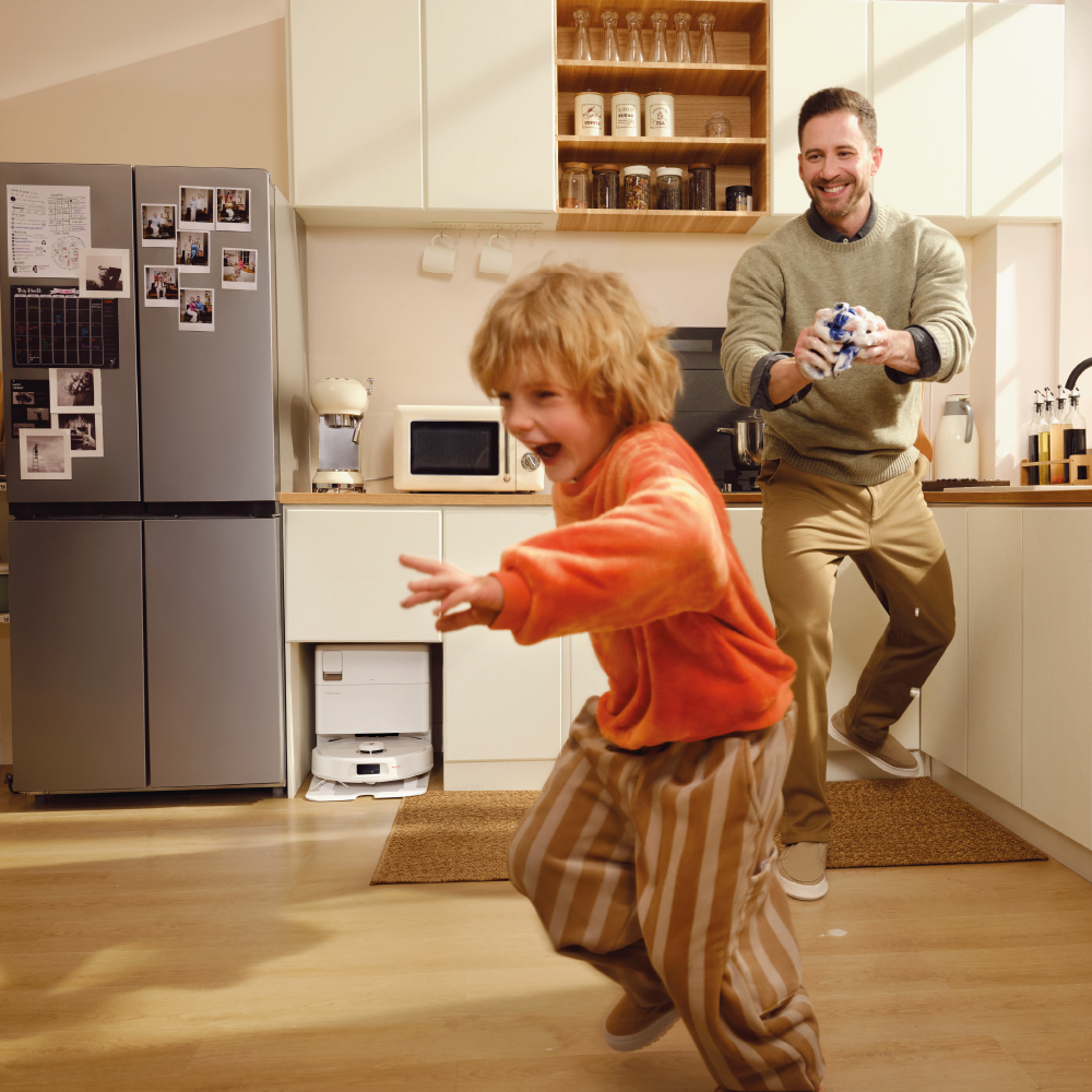 Ein Vater und ein Kind spielen in einer Küche. Das Kind rennt auf die Kamera zu. Der Vater hält ein Spielzeug. Küchengeräte und Schränke sind sichtbar.