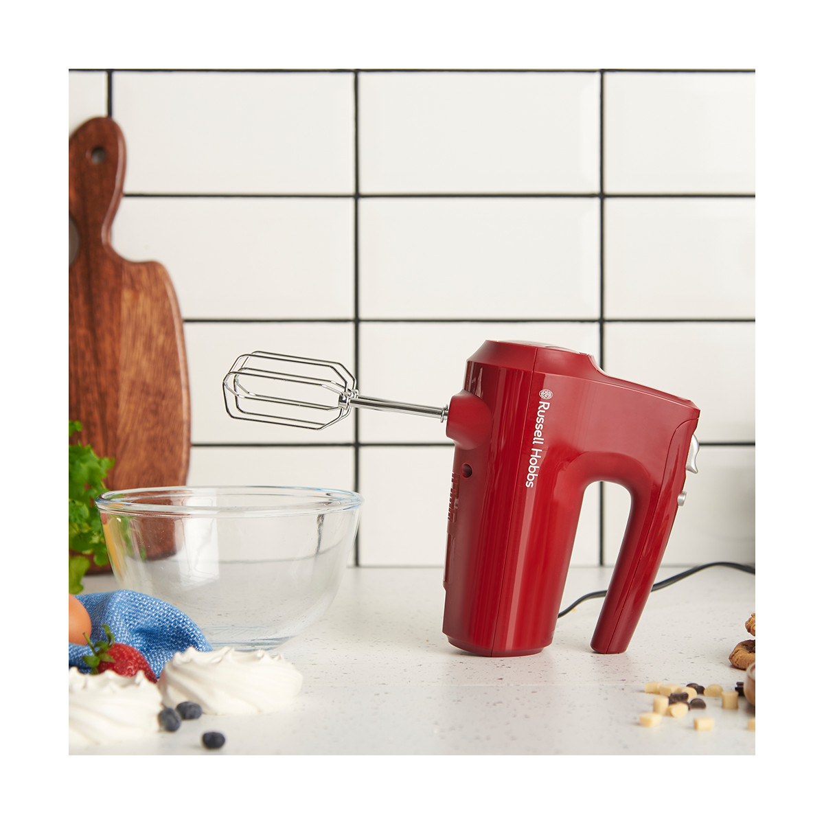 Un frullatore a mano rosso Russell Hobbs è su un bancone. Una ciotola, meringhe e un tagliere sono vicini. Parete di piastrelle bianche sullo sfondo.
