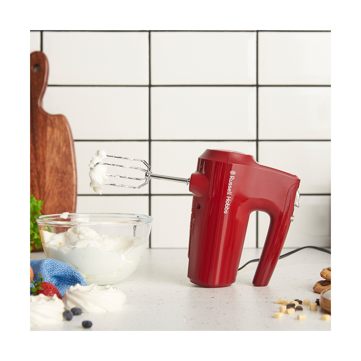 Un frullatore a mano rosso con frusta, che mescola panna bianca. Sono presenti anche una ciotola di vetro con panna, un tagliere e biscotti.