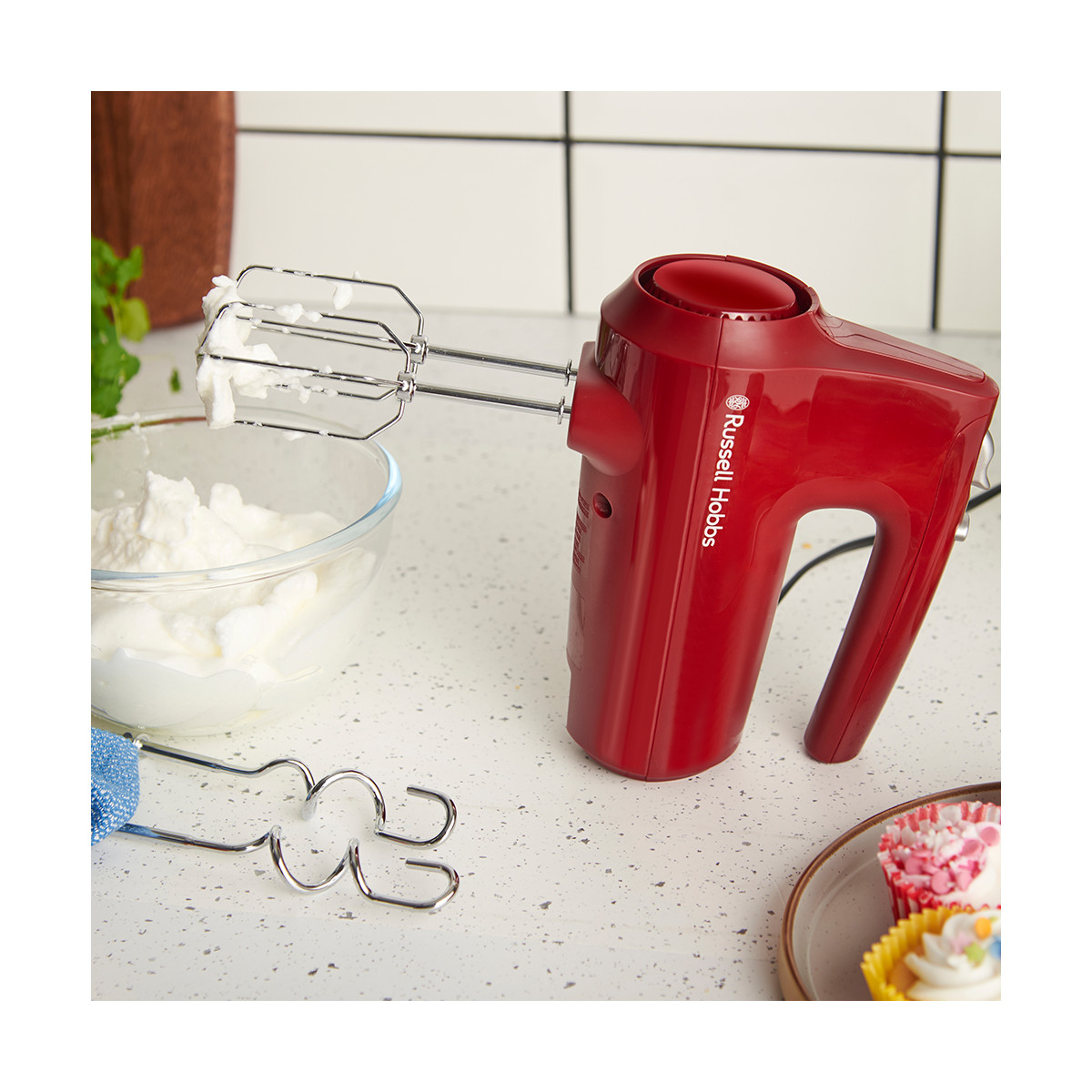 Un frullatore a mano rosso Russell Hobbs con accessori, una ciotola di panna montata e cupcake su un bancone.