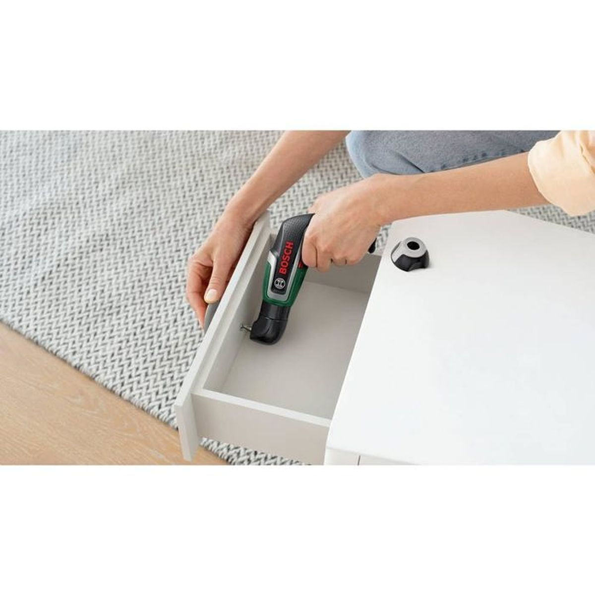 Eine Person benutzt einen Bosch-Akkuschrauber, um an einer weißen Schublade zu arbeiten. Die Schublade ist auf einem weißen Tisch geöffnet.