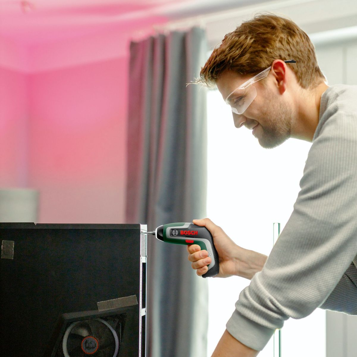 Ein Mann mit Schutzbrille benutzt einen Bosch-Schraubendreher an einem Computer. Im Hintergrund sind Vorhänge und eine rosa Wand zu sehen.