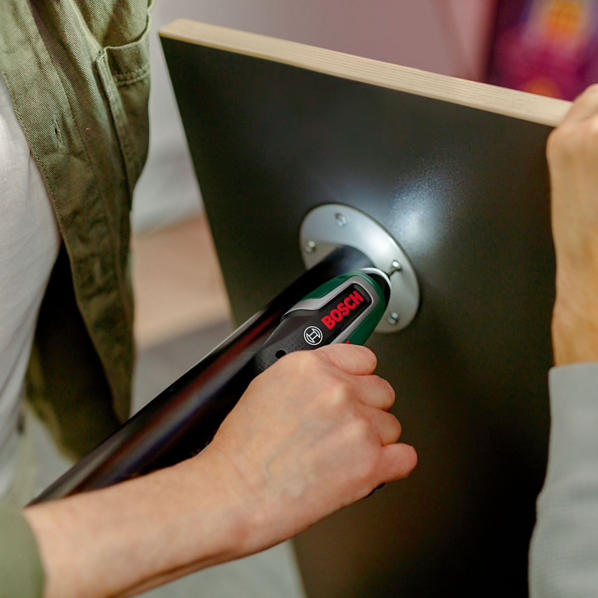 Eine Person benutzt einen Bosch-Schraubendreher, um Möbel zusammenzubauen. Der Schraubendreher ist grün und schwarz. Das Möbelstück ist schwarz.