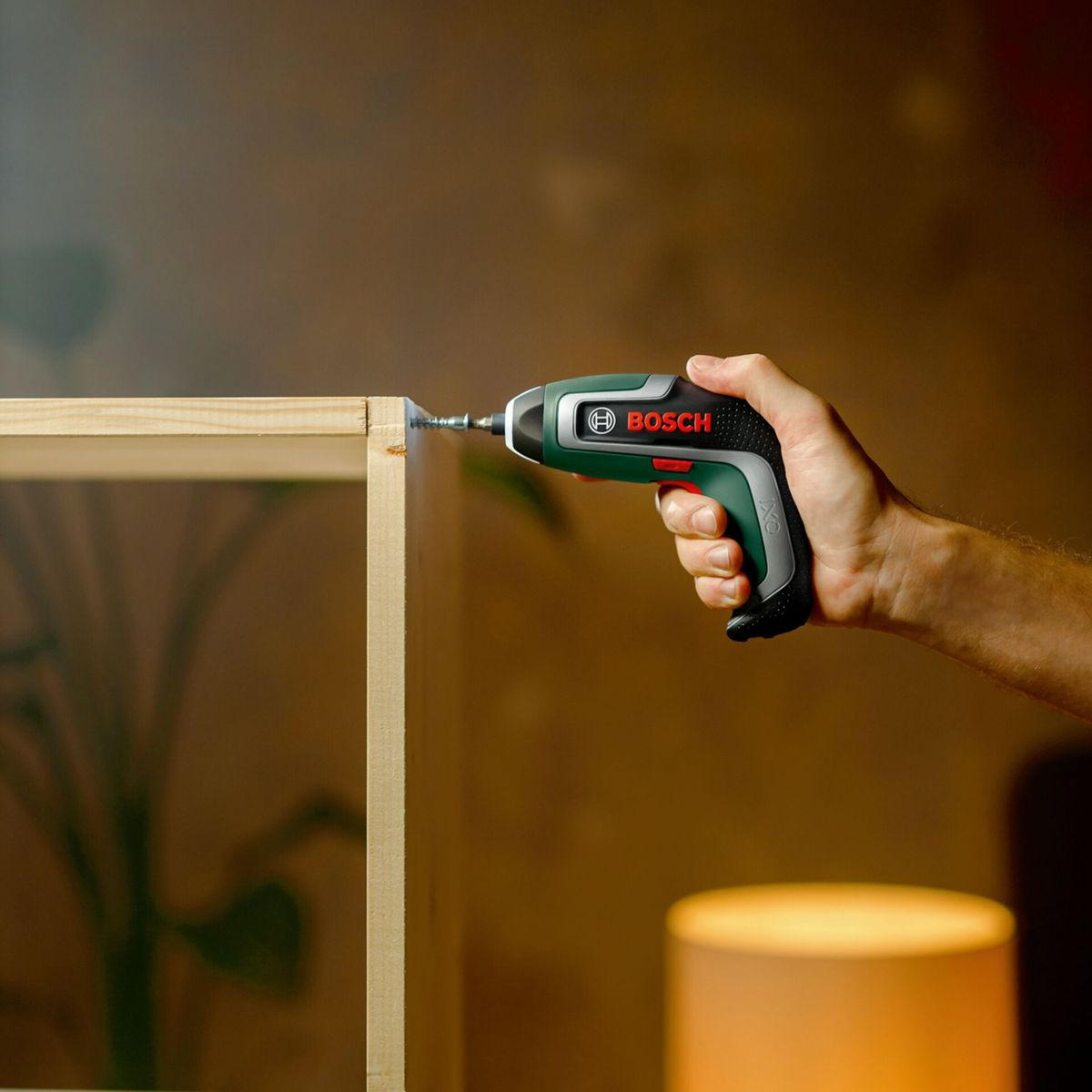 Eine Hand benutzt einen grünen und schwarzen Bosch-Schraubendreher, um eine Schraube in eine Holzkonstruktion zu schrauben, drinnen.