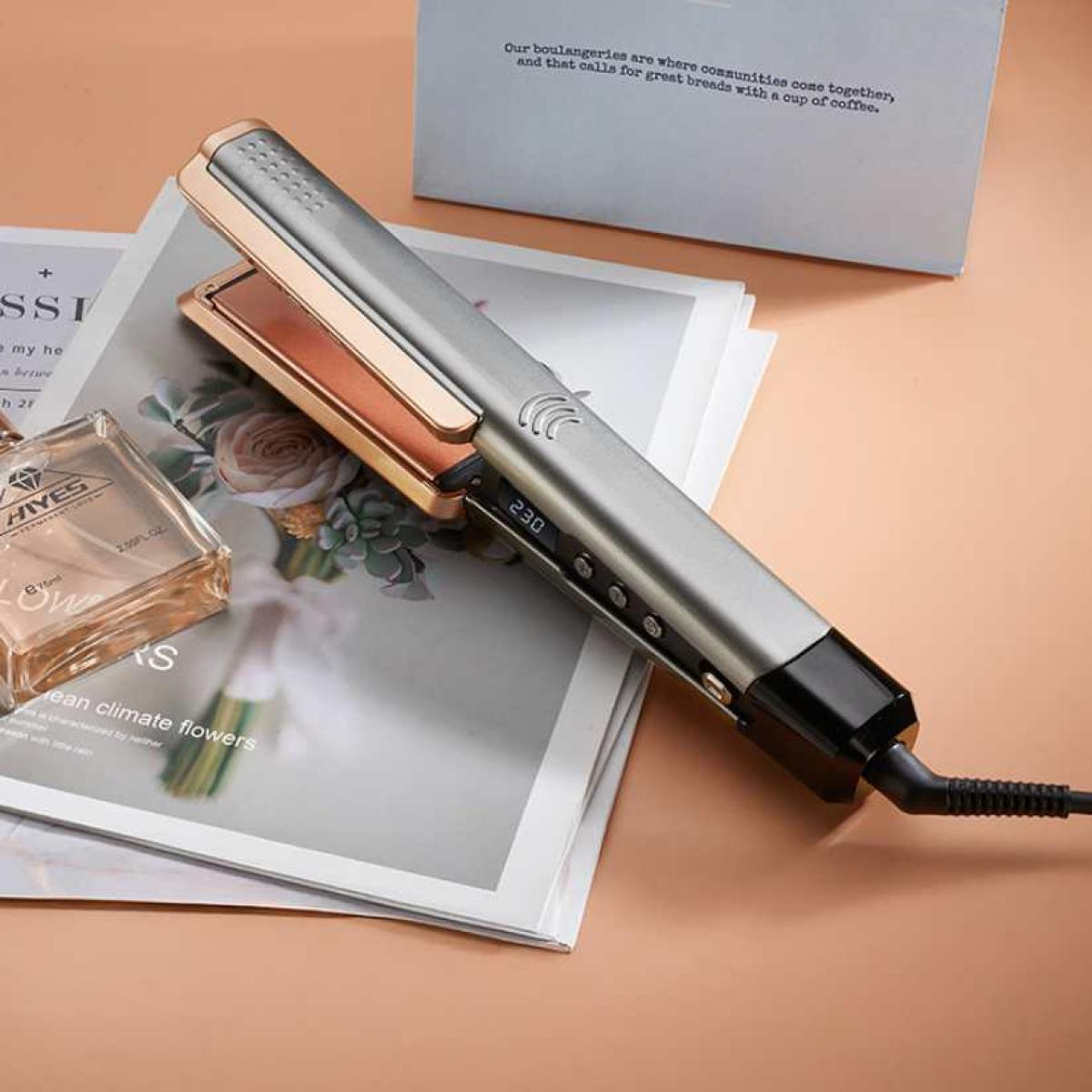 Silberner Haarglätter mit roségoldenen Platten auf einem Magazin. Parfümflasche und eine Schachtel im Hintergrund.