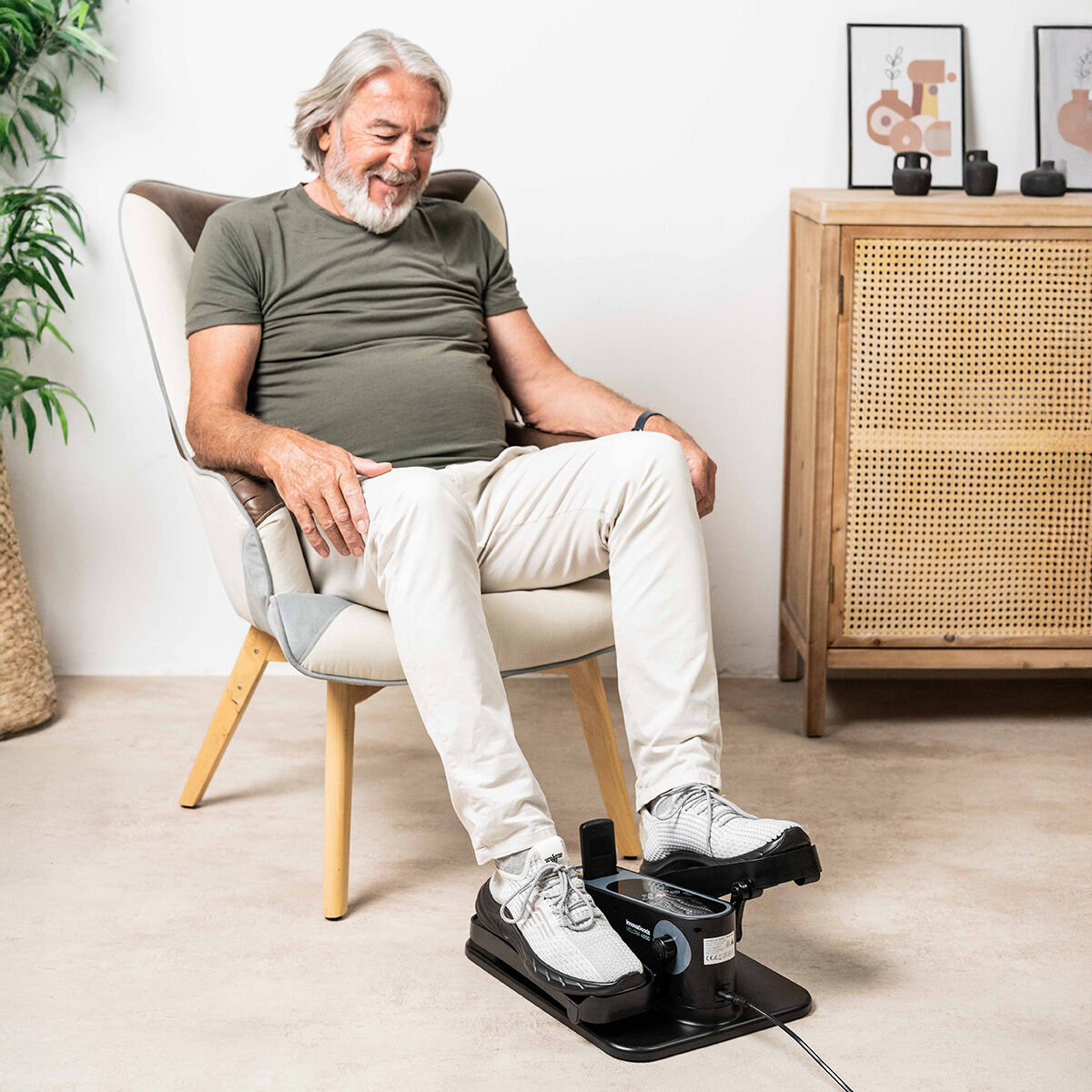 Un homme âgé utilise un pédalier assis. Il porte une chemise verte et un pantalon blanc. Le cadre est un salon.