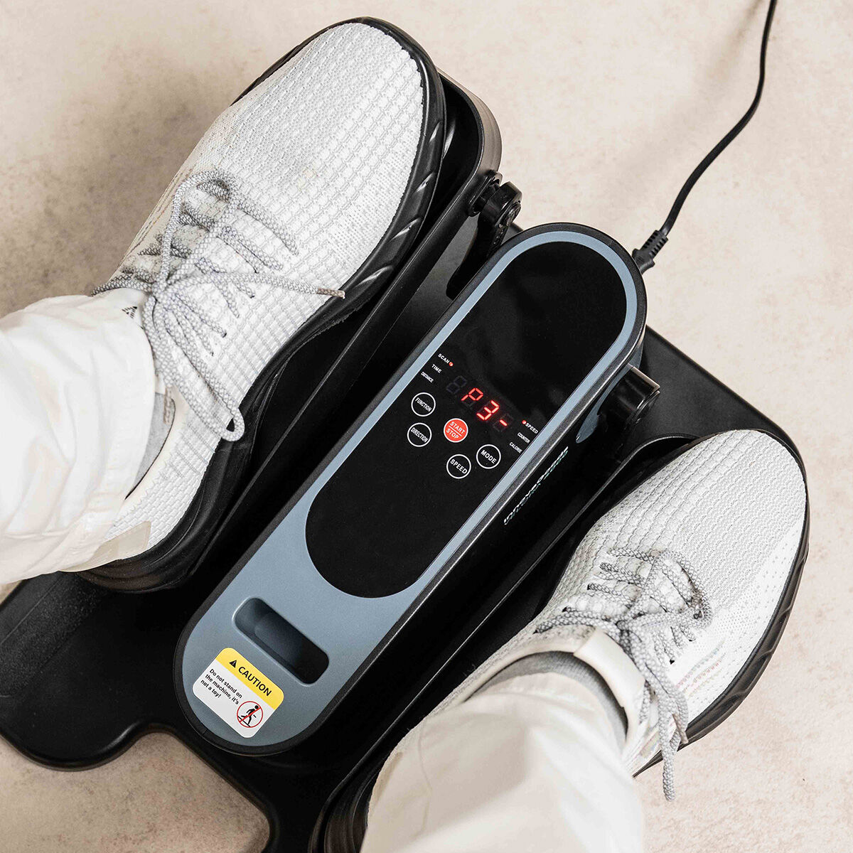 Pieds d'une personne sur un stepper. Baskets blanches sur pédales noires. Un écran numérique affiche des chiffres. Un autocollant de précaution est visible.