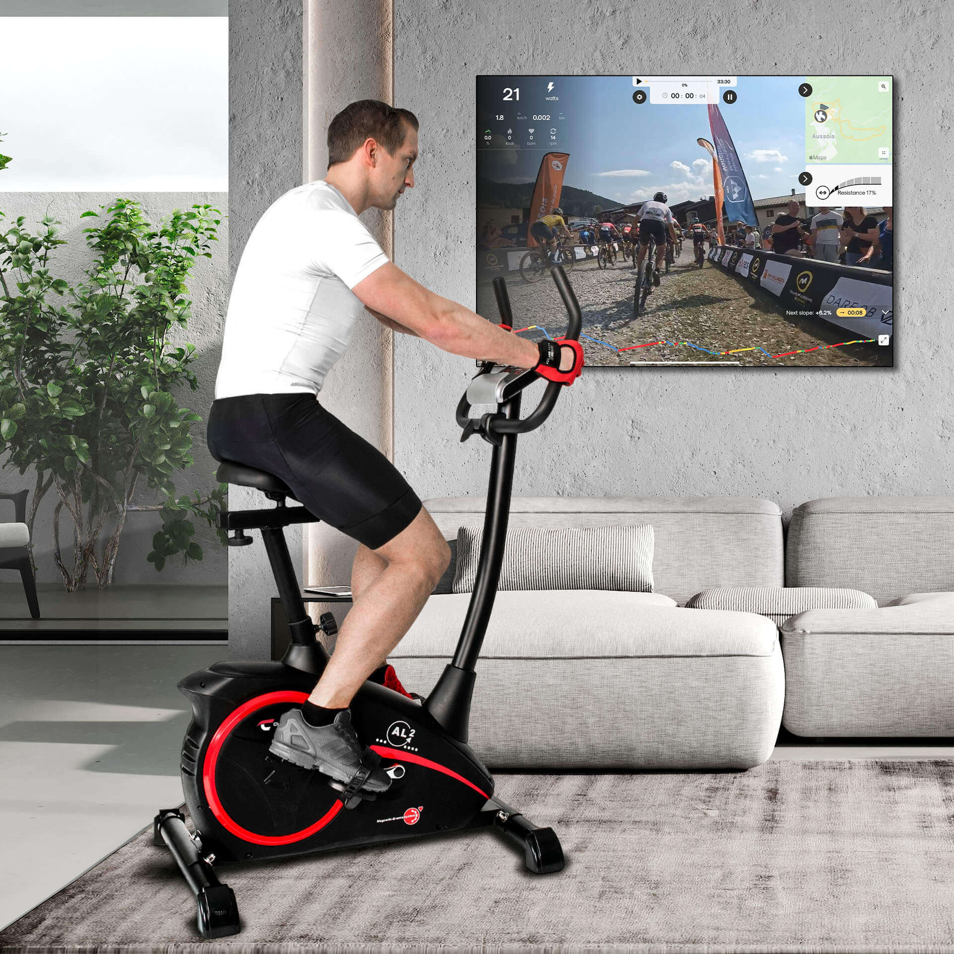 Ein Mann trainiert auf einem Heimtrainer und schaut sich ein Rennen auf einem Bildschirm an. Das Fahrrad ist schwarz und rot, drinnen.