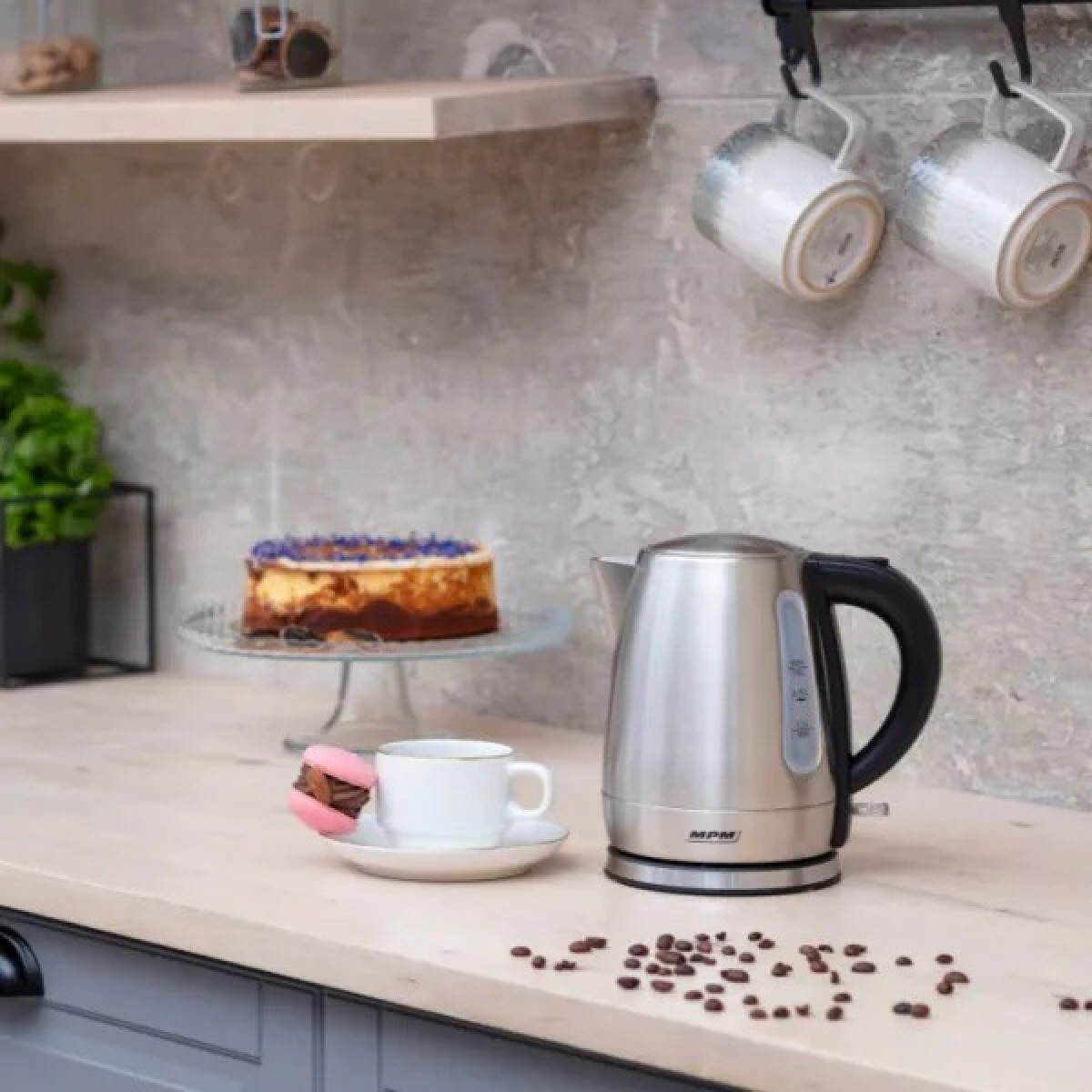 Scena cucina con bollitore, torta, tazza e macaron. Chicchi di caffè sul bancone. Tazze appese sullo sfondo.