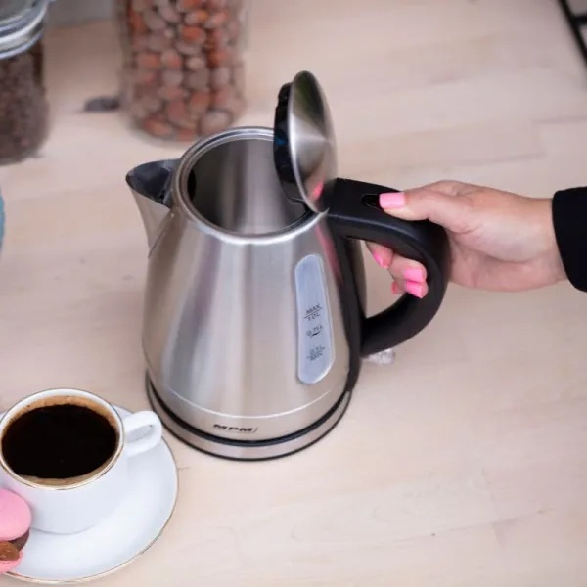 Un bollitore in acciaio inossidabile con coperchio aperto, tenuto da una mano con unghie rosa. Una tazza di caffè e barattoli sullo sfondo.