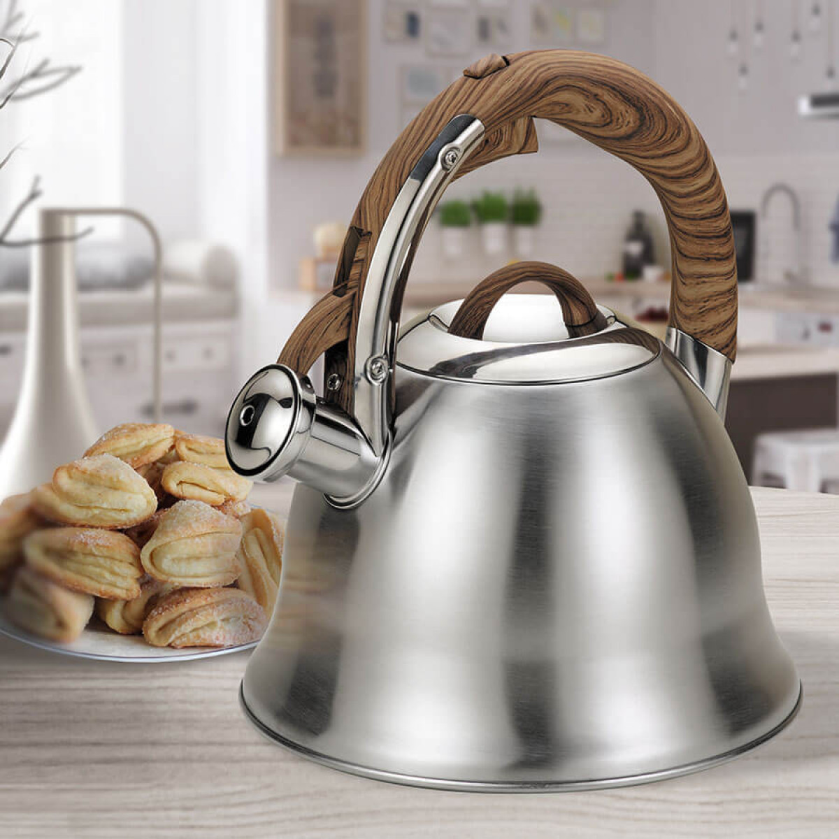 Una teiera in acciaio inossidabile con manico in legno e un piatto di biscotti su un tavolo di legno. Cucina sullo sfondo.