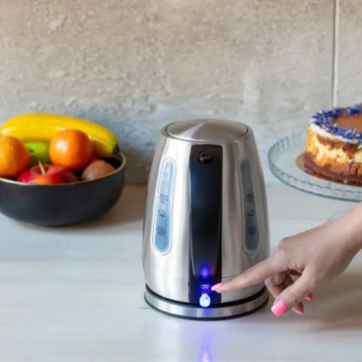 Una mano preme il pulsante di accensione di un bollitore elettrico argentato. Una ciotola di frutta e una torta sullo sfondo.