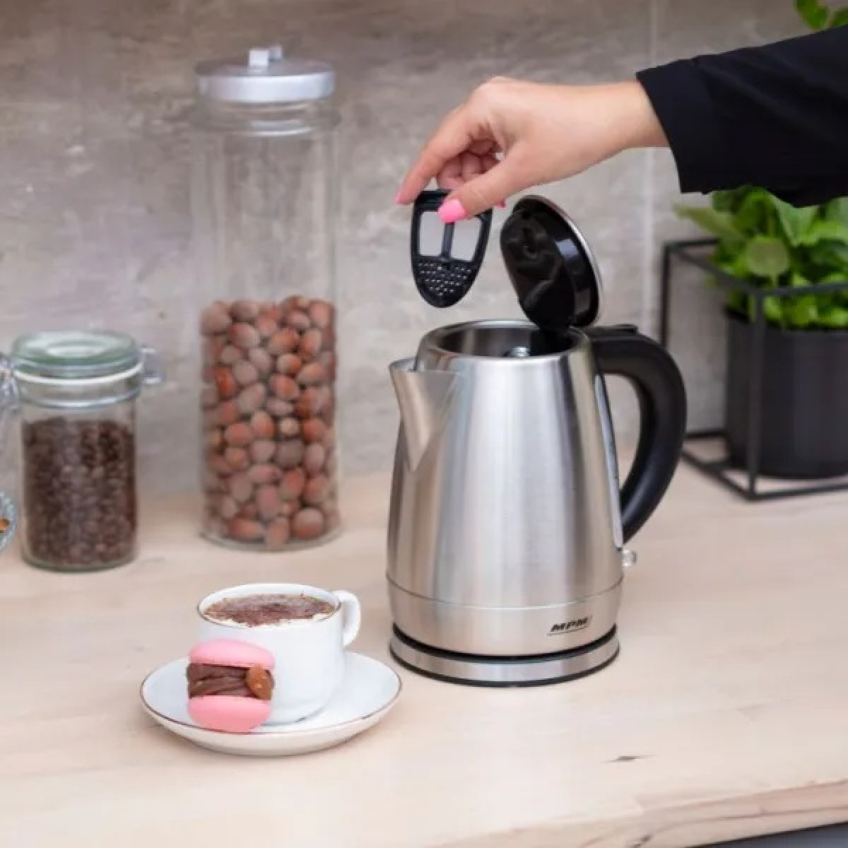Una mano mette un filtro in un bollitore elettrico argentato. Una tazza di caffè e un macaron rosa sono su un tavolo. Barattoli di noci sullo sfondo.