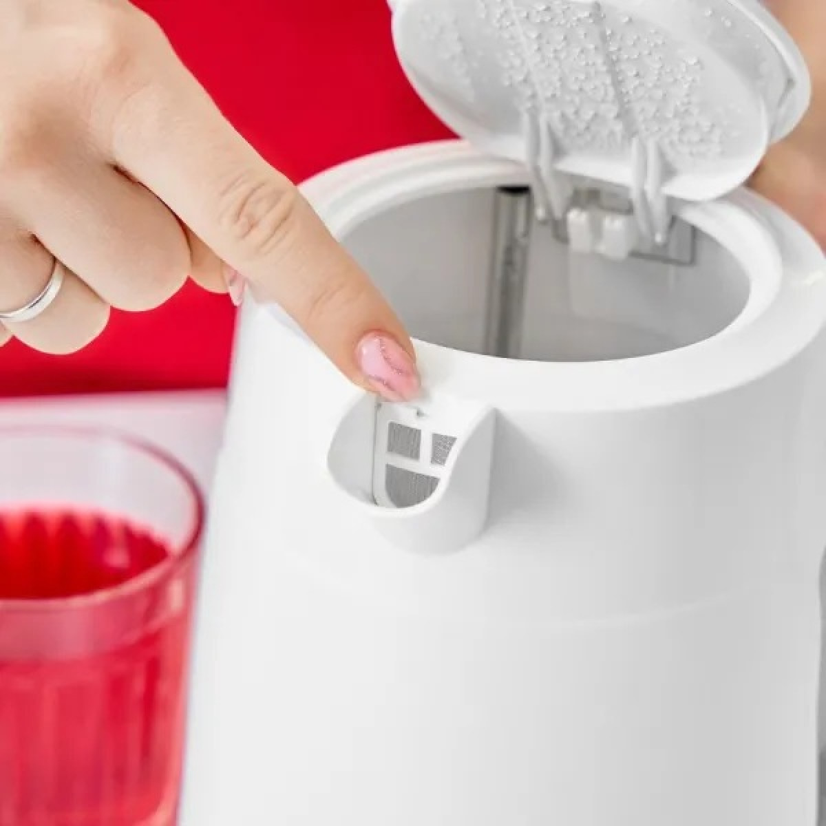 Una mano indica un bollitore elettrico bianco con coperchio aperto. Un bicchiere di liquido rosso in primo piano.