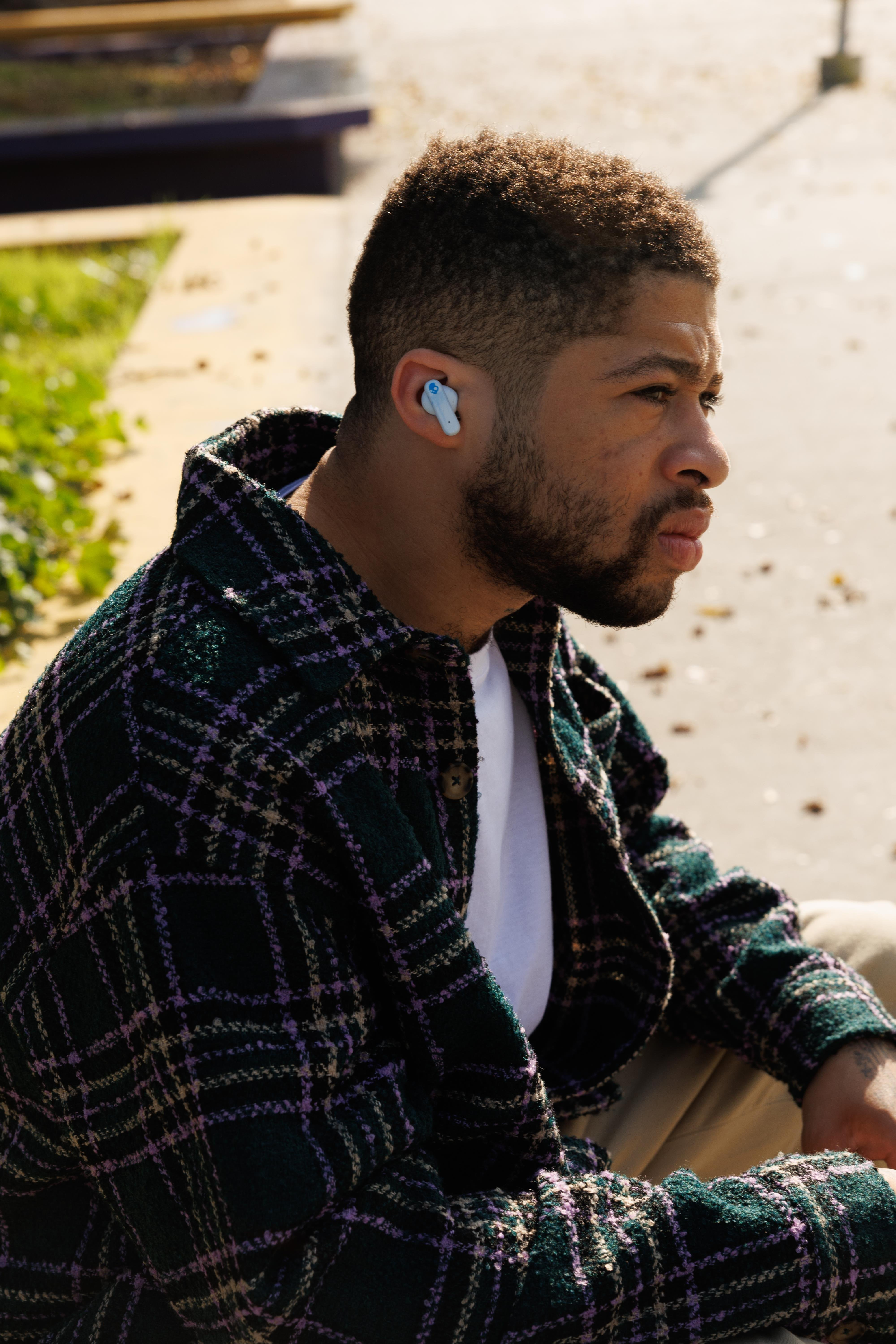 Mężczyzna z brodą w kratowanej kurtce i niebieskiej słuchawce. Siedzi na zewnątrz w białej koszulce.