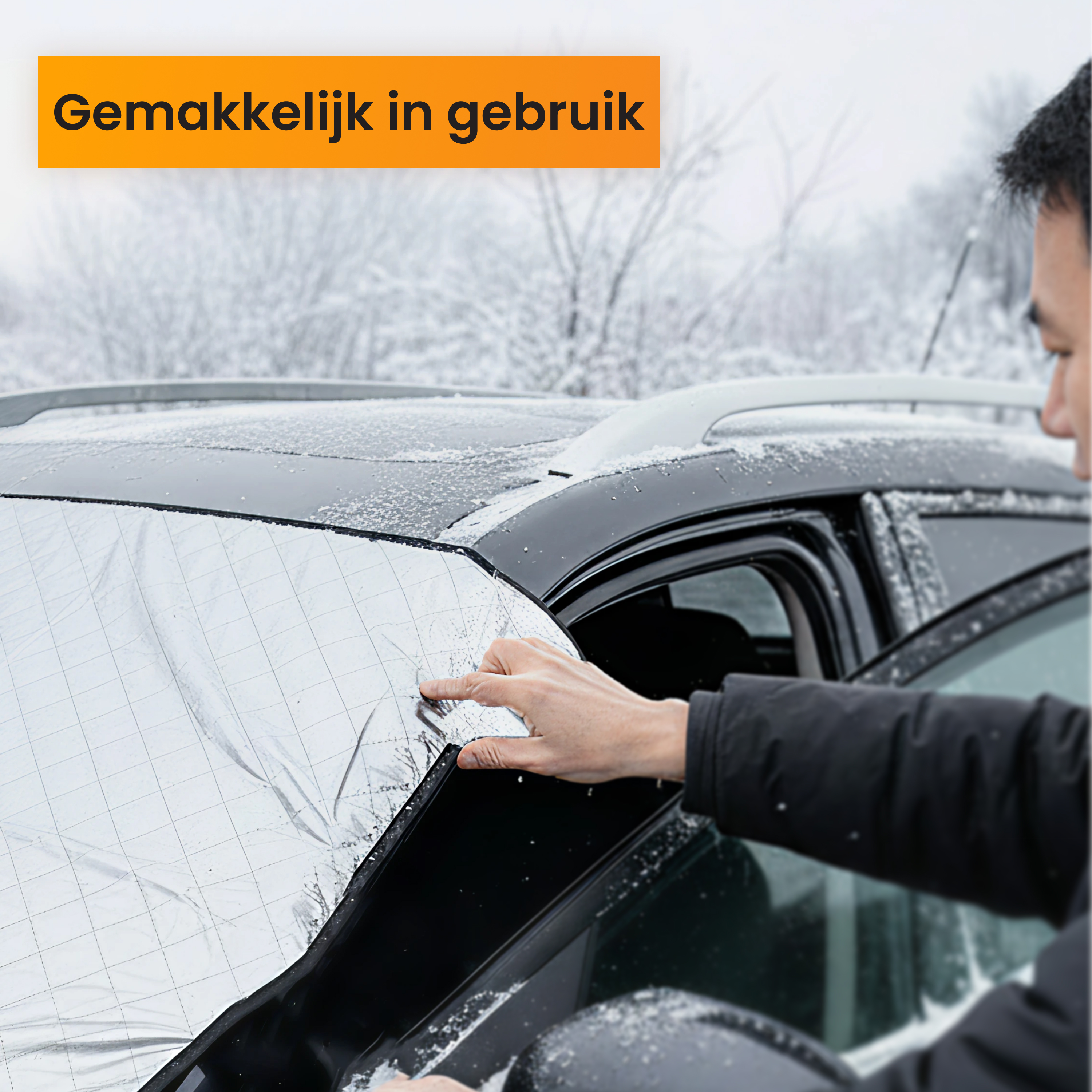 A person installing a silver windshield cover on a car in a snowy environment. The cover has a grid pattern.