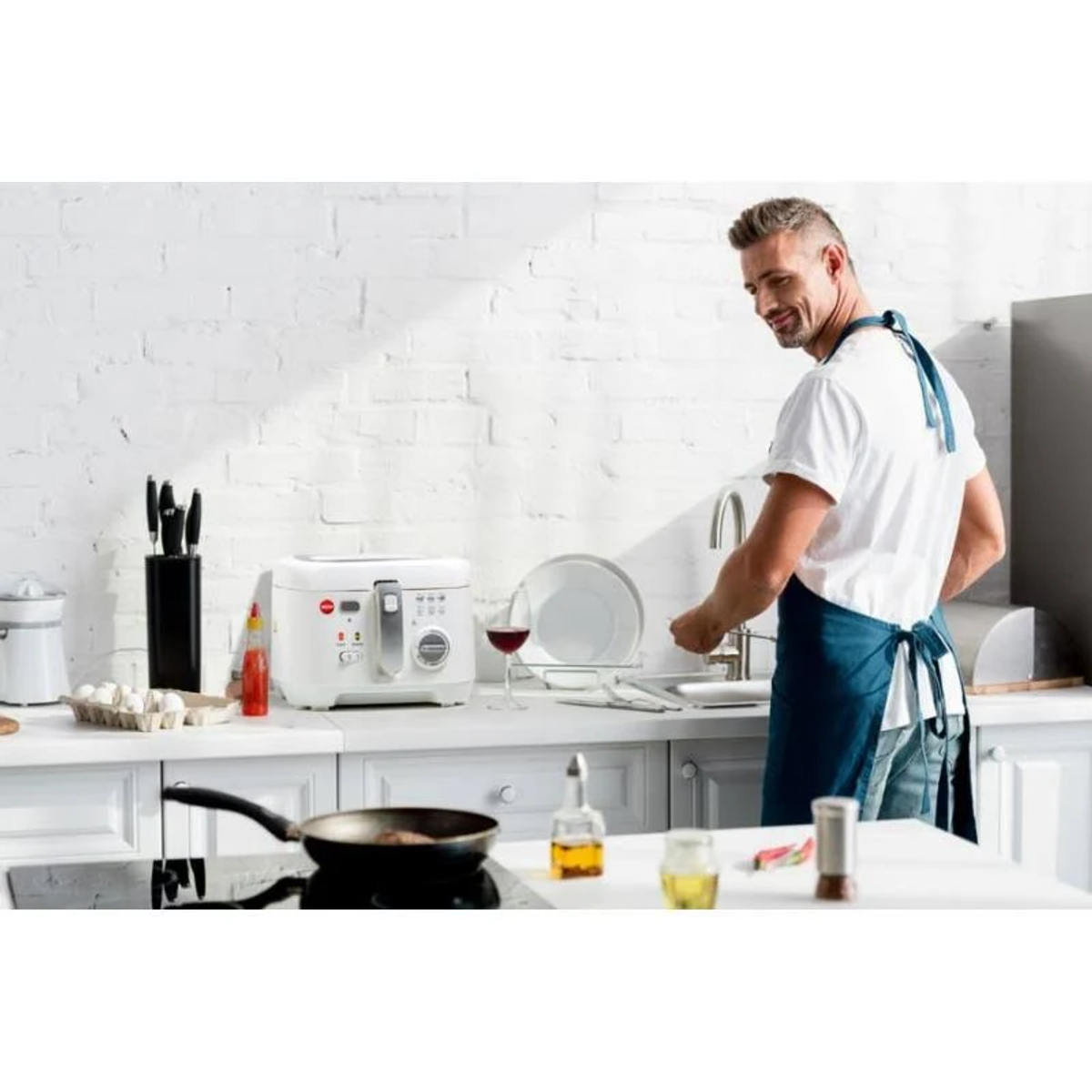 Mężczyzna w kuchni, zmywa naczynia. Na blacie frytownica, patelnia z jedzeniem, kieliszek do wina i butelki z olejem.