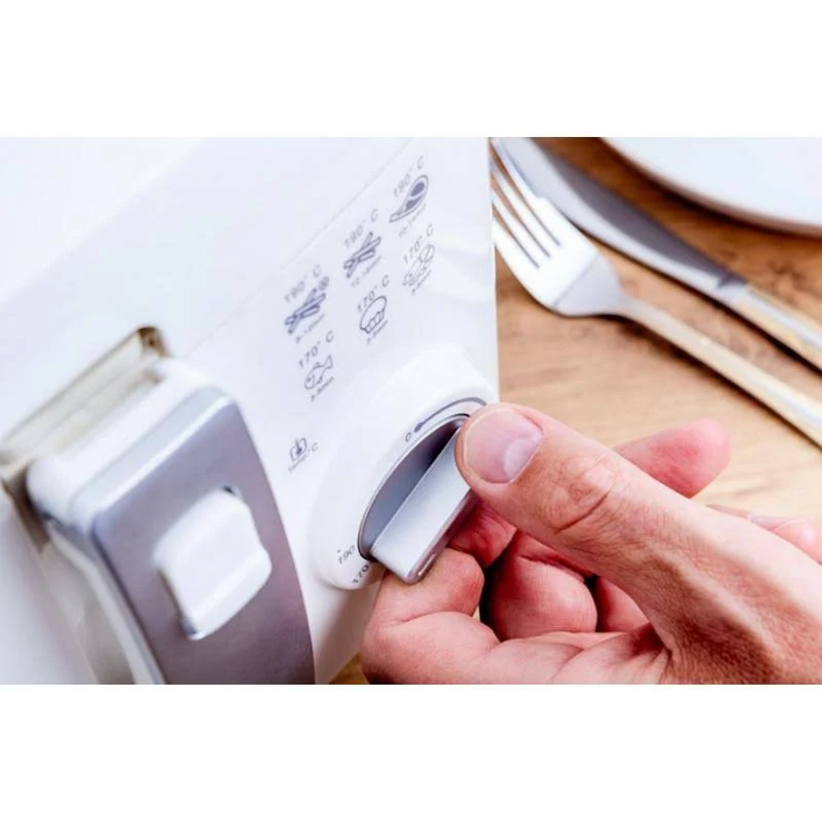 Ręka obraca pokrętło białego urządzenia. Widelec i nóż leżą na drewnie, w tle biały talerz.