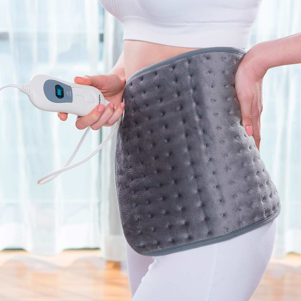 Kobieta w bieli trzyma szarą poduszkę rozgrzewającą na plecach. W drugiej ręce trzyma kontroler.