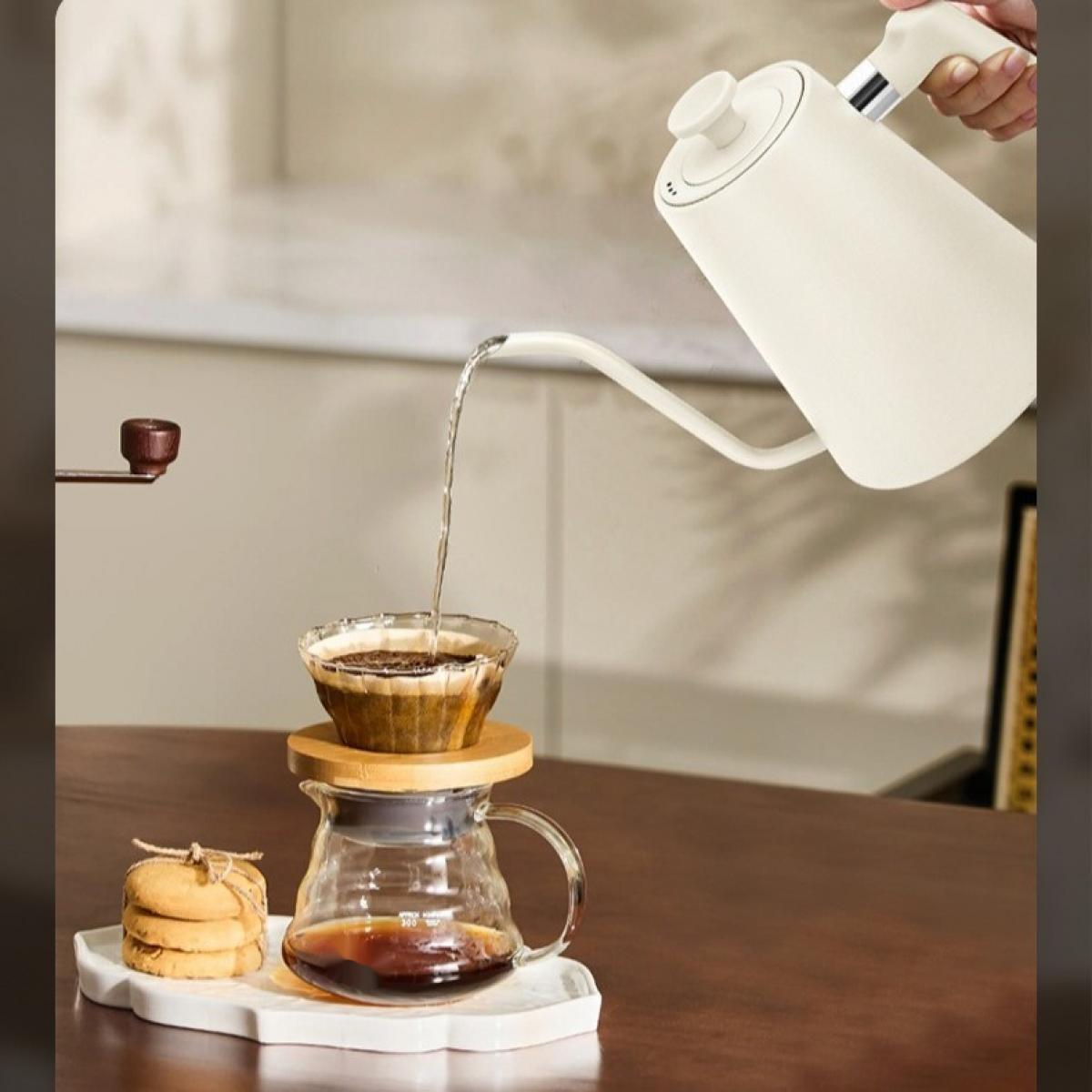 Eine Person gießt Wasser aus einem weißen Wasserkocher in einen Kaffeefilter. Eine Glaskaraffe und Kekse stehen auf dem Tisch.