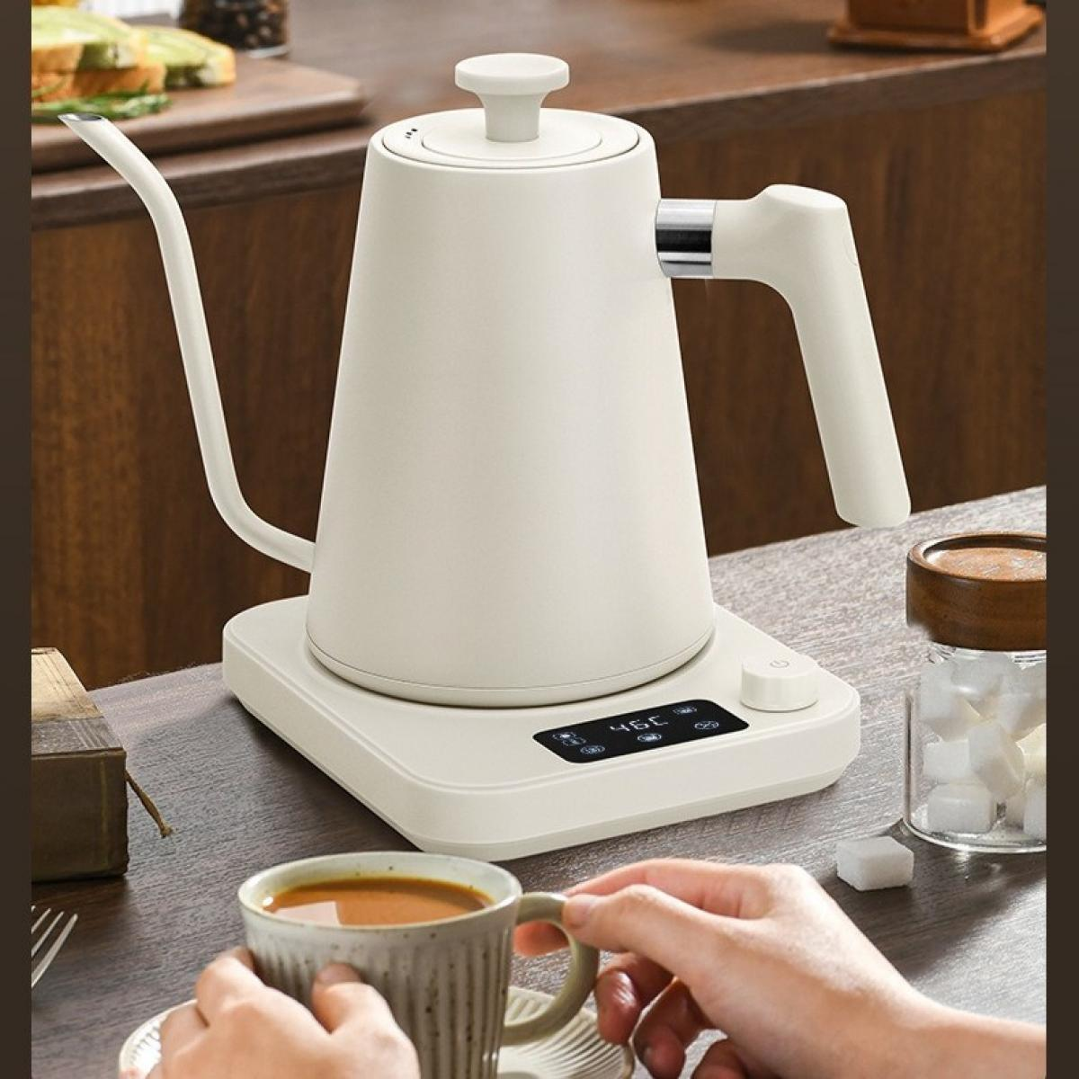 Ein cremefarbener Wasserkocher steht auf einer digitalen Basis. Eine Person hält eine Kaffeetasse. Zuckerwürfel und ein Glas stehen auf dem Tisch.