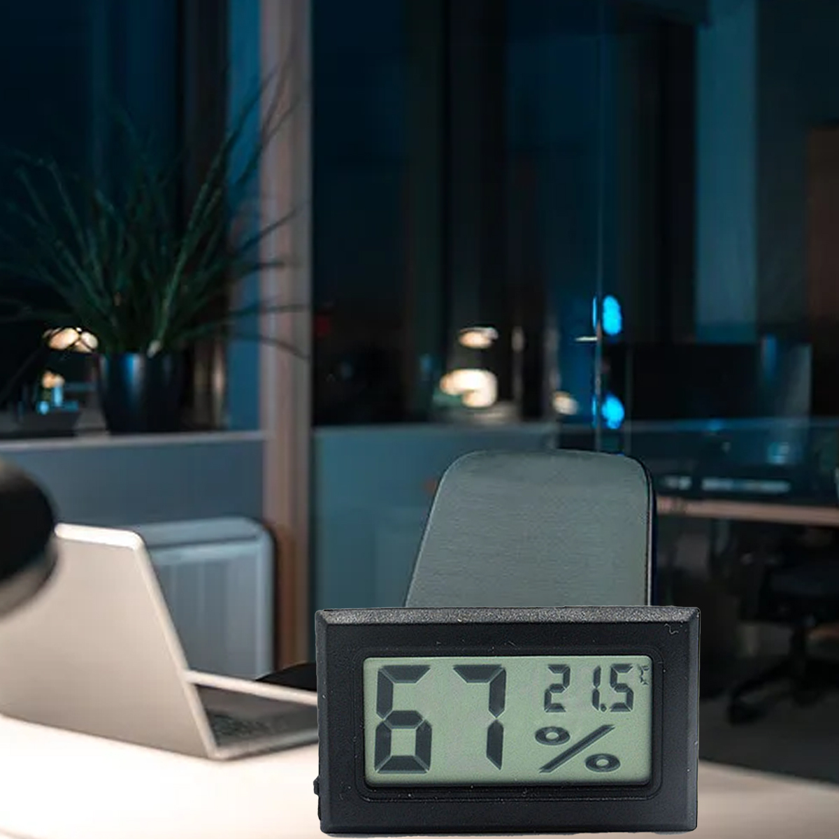 Ein digitales Thermometer zeigt 67 % Luftfeuchtigkeit und 21,5 Grad an. Auf einem Schreibtisch sind ein Laptop und eine Pflanze zu sehen, Büro im Hintergrund.