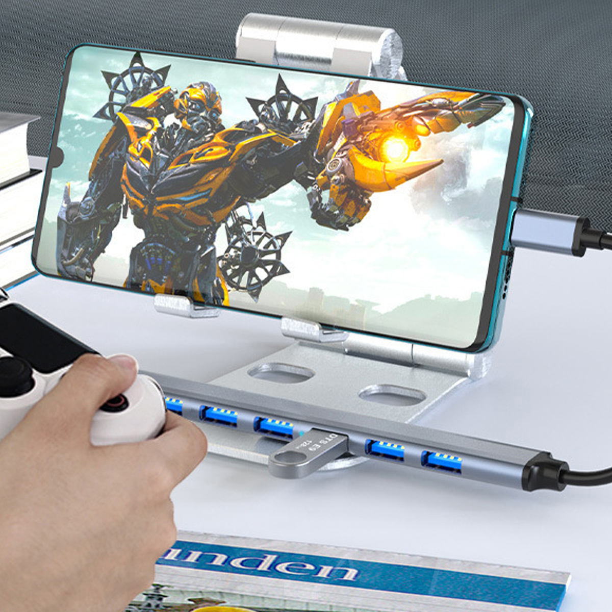 Ein Telefon mit Roboterbild, an ein Ladegerät angeschlossen. Eine Hand hält einen Controller. Ein USB-Hub im Vordergrund.