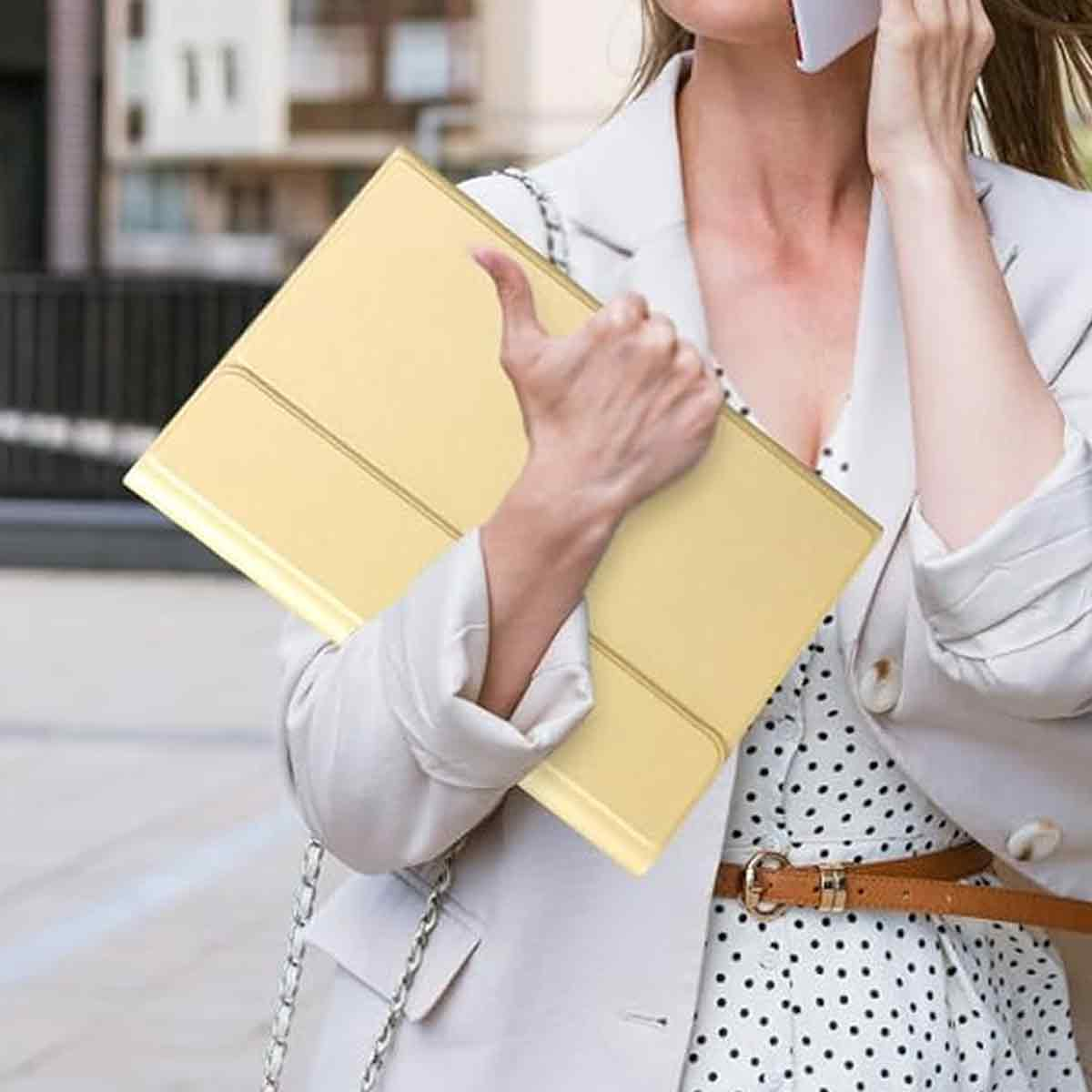 Eine Frau hält eine gelbe Mappe und telefoniert. Sie trägt einen weißen Blazer über einem gepunkteten Kleid und einen braunen Gürtel.