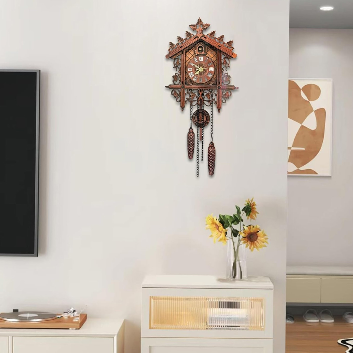 Eine Kuckucksuhr hängt an einer weißen Wand. Darunter steht ein weißer Schrank mit Sonnenblumen in einer Vase. Ein Fernseher und Kunstwerke sind ebenfalls zu sehen.