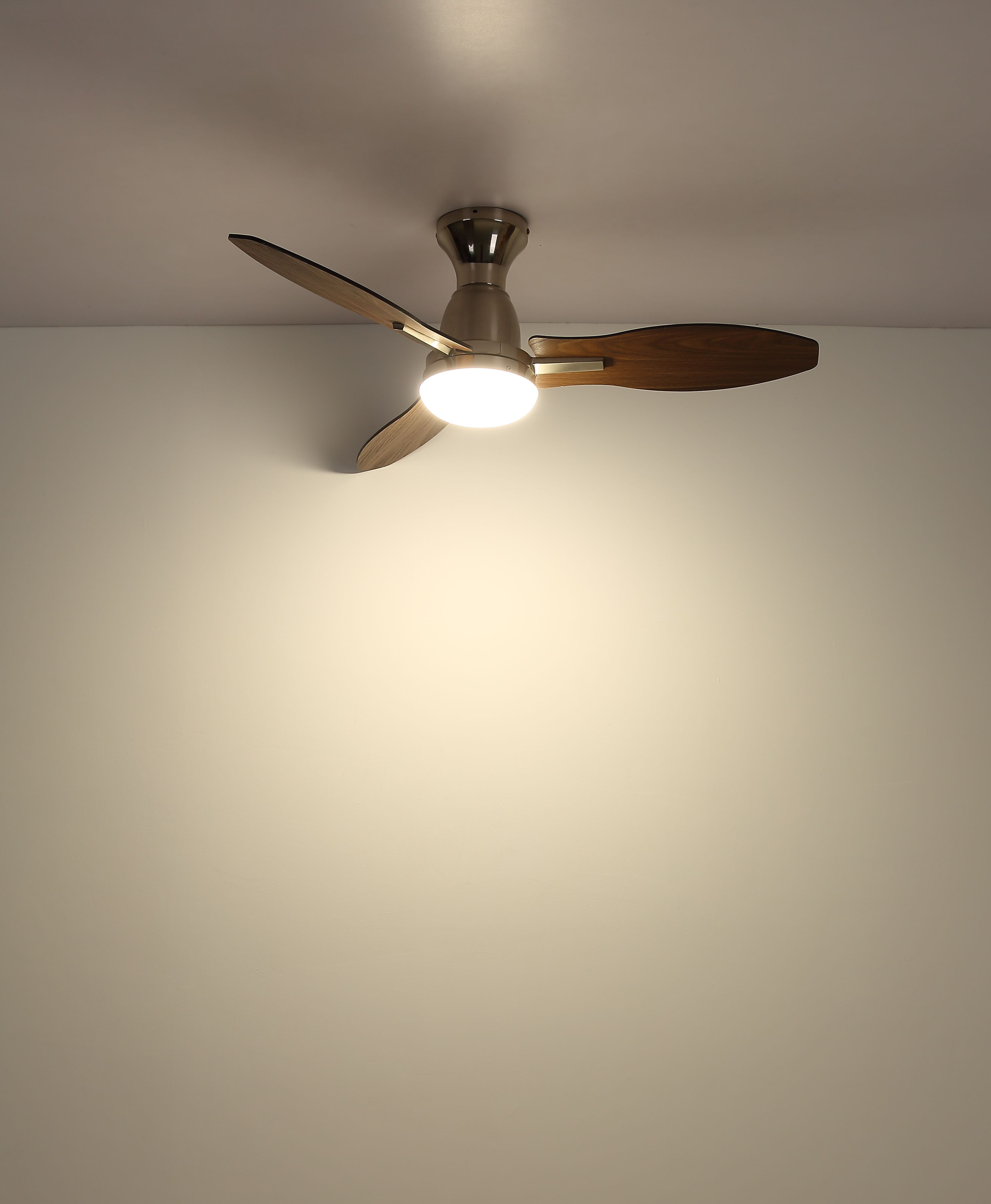 Ein Deckenventilator mit drei Holzflügeln und einer zentralen Leuchte ist an einer weißen Decke montiert.