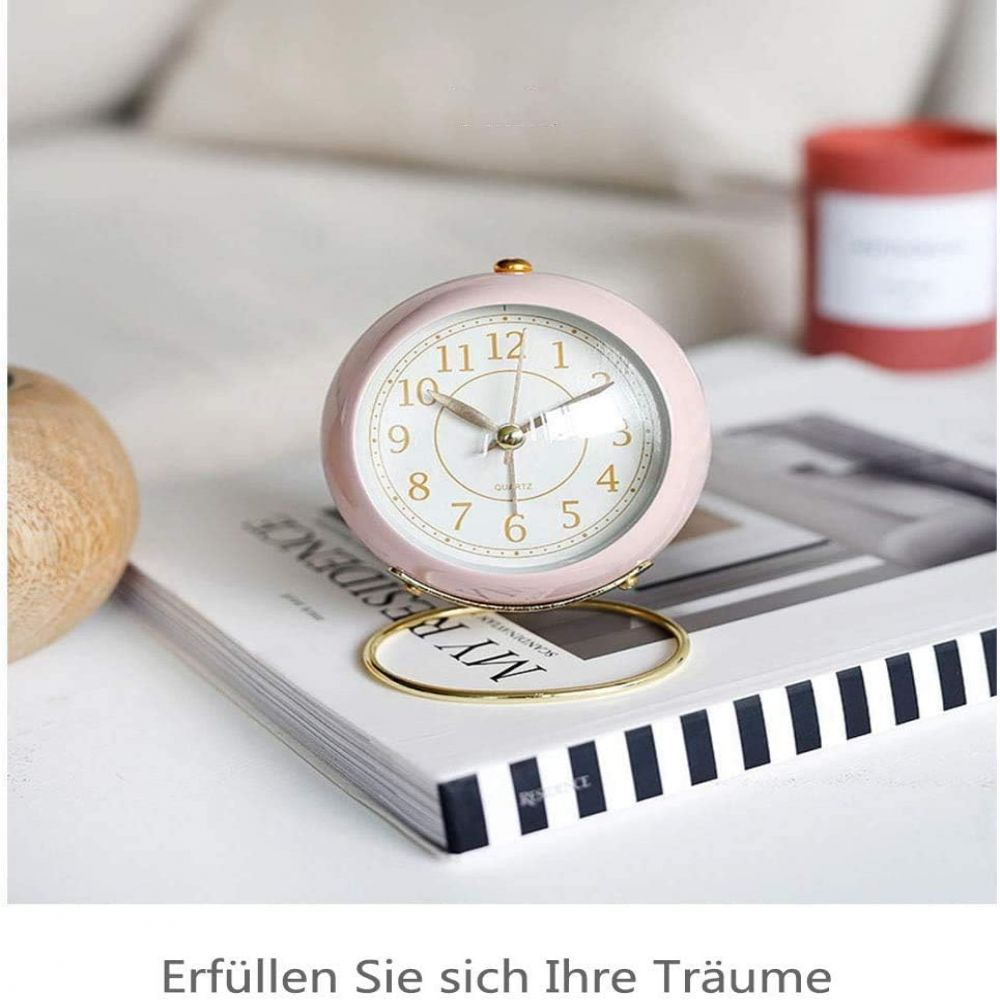 Ein rosa Quarzwecker mit goldenen Zeigern und Zahlen steht auf einem Buch. Eine Holzkugel und eine rote Kerze sind im Hintergrund.