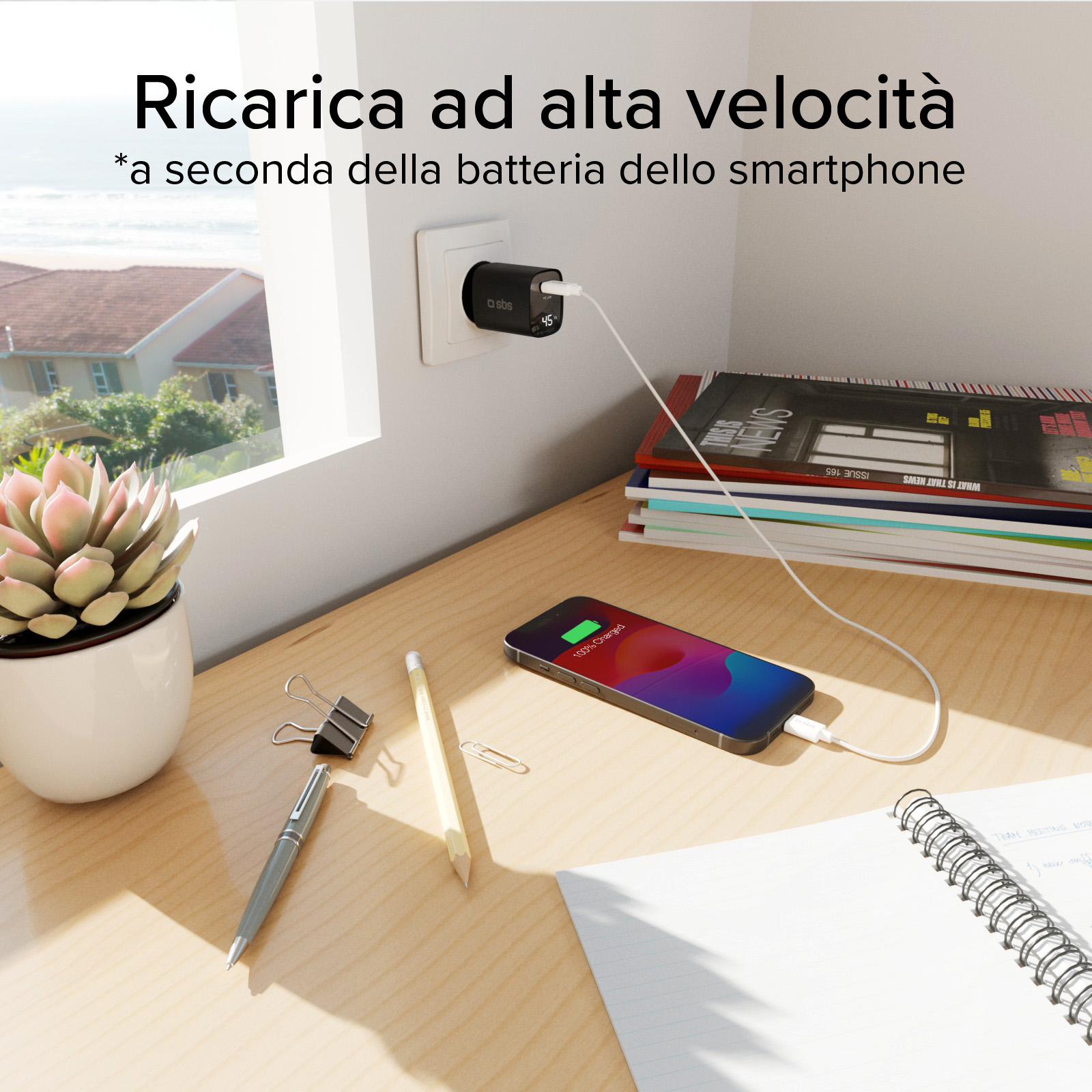 Telefon ładuje się na drewnianym biurku. Ładowarka jest podłączona do gniazdka ściennego. Sukulent stoi w doniczce. Czasopisma są ułożone w stos.