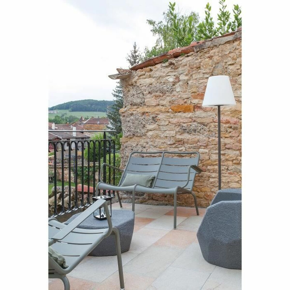 Terrasse mit grauen Metallmöbeln, Steinmauer und Lampe. Blick auf ein Dorf und grüne Hügel unter bewölktem Himmel.