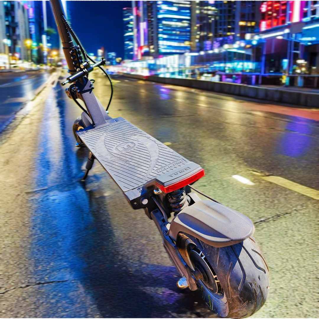 Hulajnoga elektryczna na mokrej drodze w nocy. Światła miasta w tle. Hulajnoga jest czarna i szara.