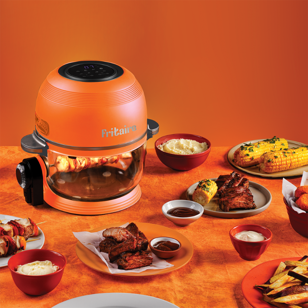 Oranger Luftfritteuse mit Drehspieß, der verschiedene Lebensmittel wie Rippchen und Mais zubereitet. Gerichte mit Saucen, Pommes und Dips stehen auf einem orangen Tisch.