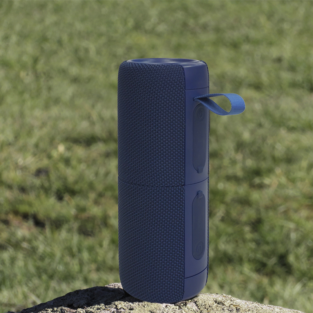 Granatowy głośnik przenośny stoi na skale, na tle rozmytej zielonej trawy. Ma uchwyt.