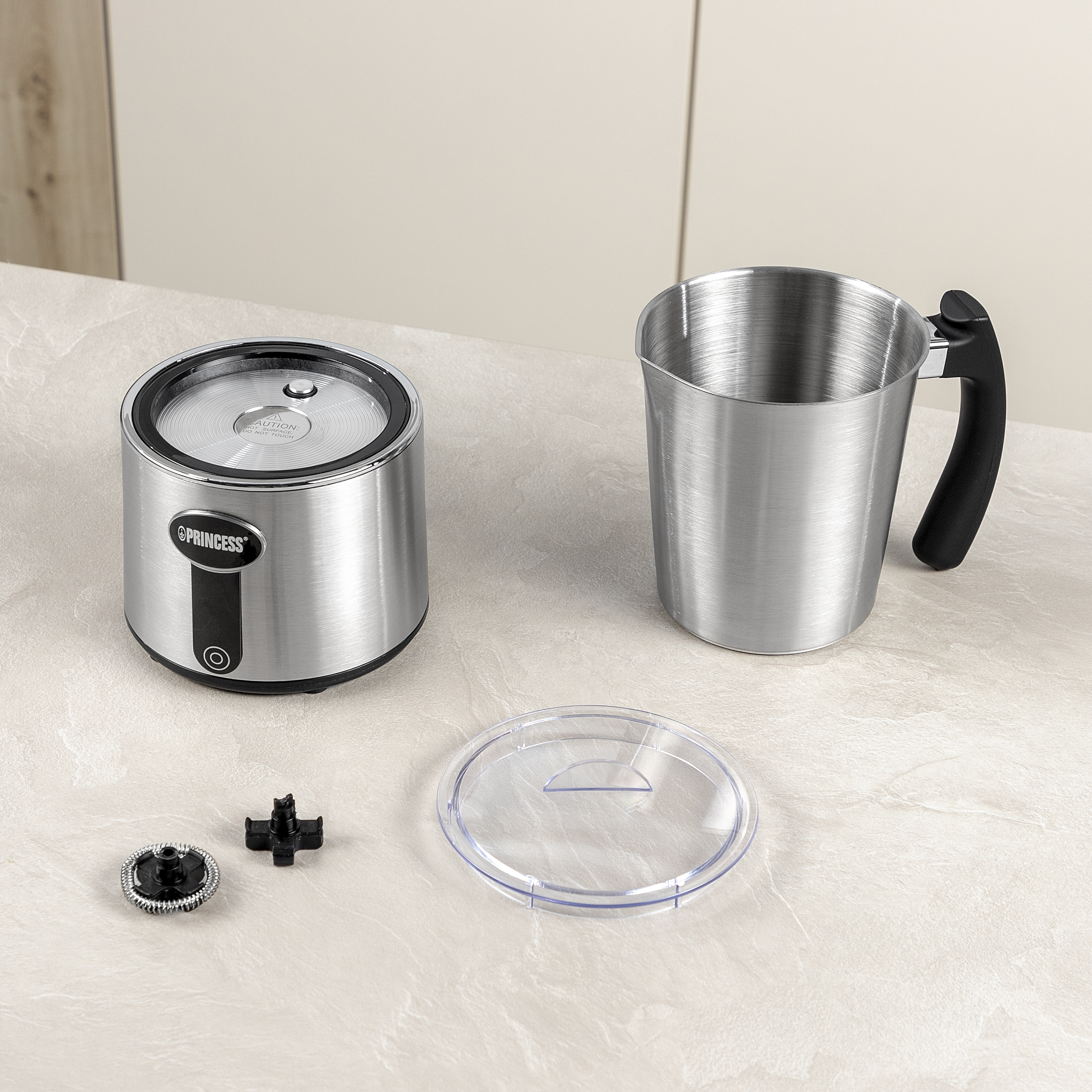 Srebrny spieniacz do mleka z częściami na jasnym blacie. Zawiera dzbanek do spieniania, pokrywkę i akcesoria. Tło jest neutralne.