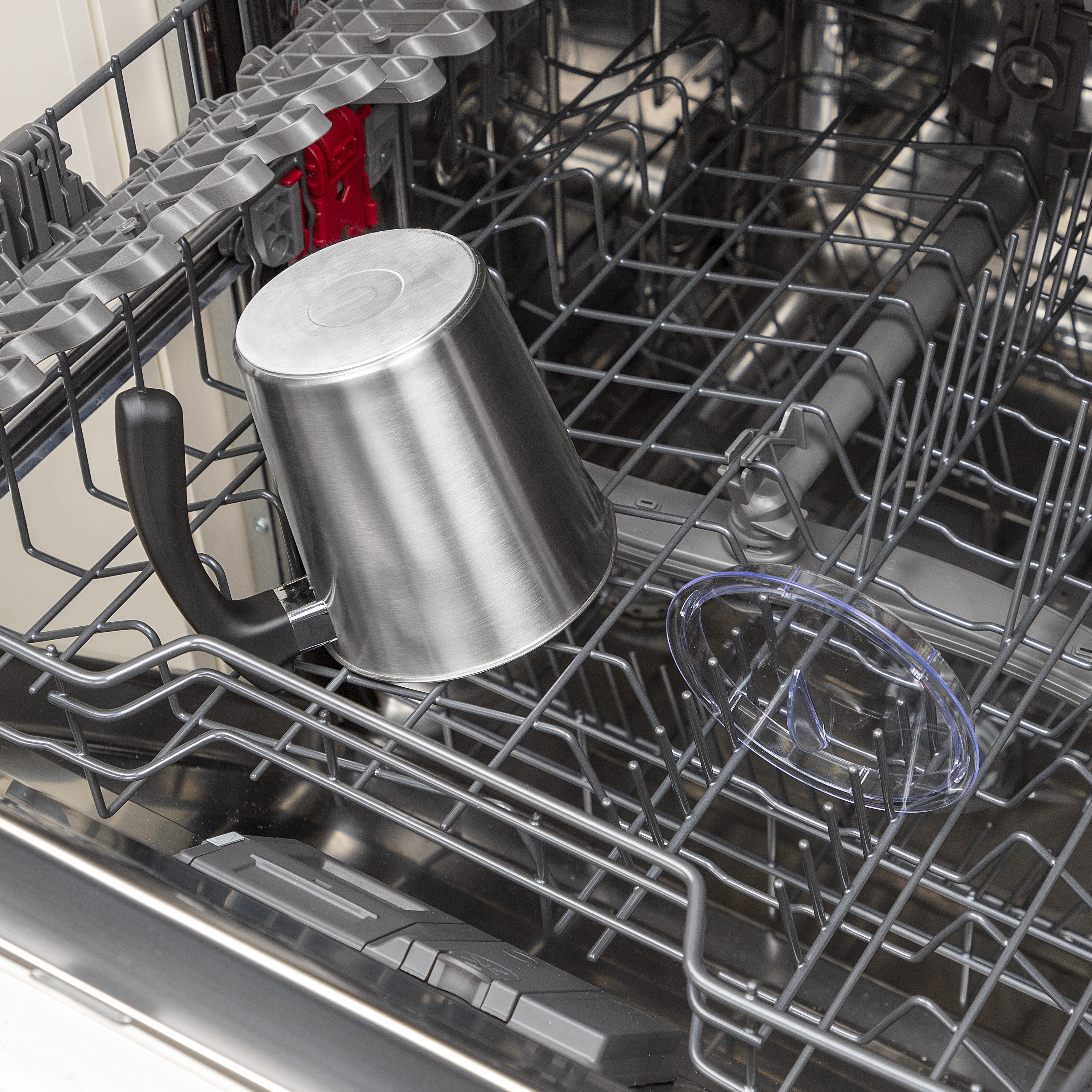Kubek ze stali nierdzewnej znajduje się w zmywarce. Widoczna jest również przezroczysta plastikowa pokrywa i metalowe ramy.
