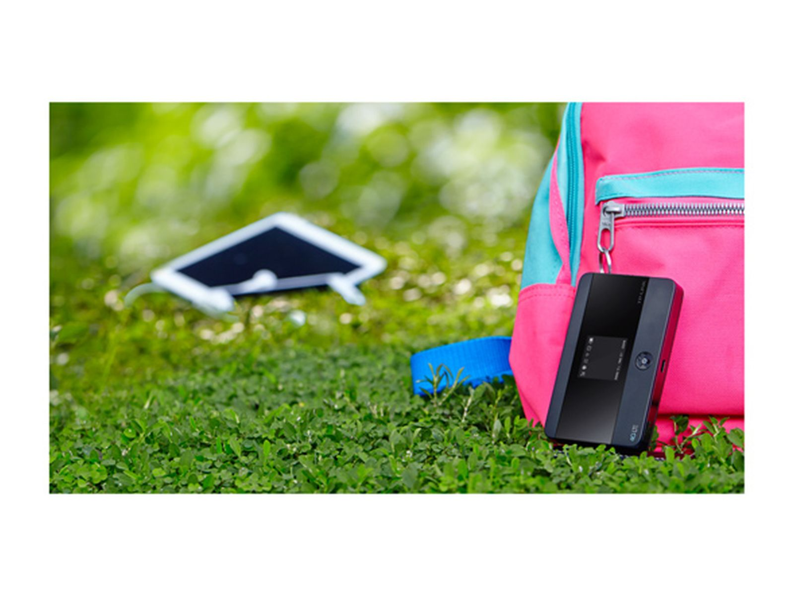Siyah bir cihaz ve mavi detaylı pembe bir sırt çantası çimenlerin üzerinde duruyor. Arka planda bir tablet ve mavi bir kayış, bulanık yeşil bir arka plan var.